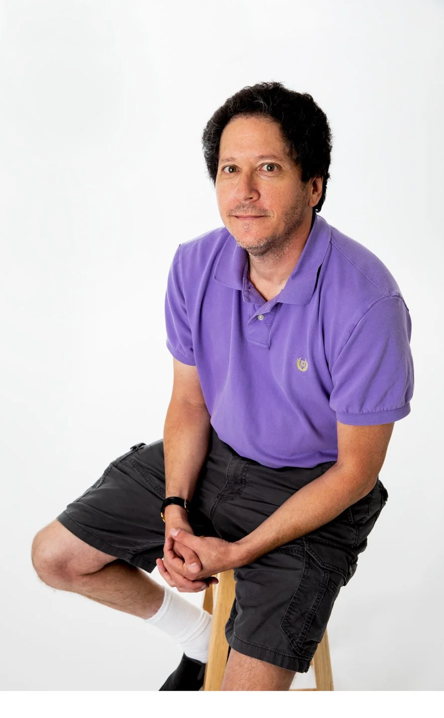 A man sitting on a wooden stool against a plain white background, wearing a purple polo shirt, black shorts, and white socks, looking at the camera.