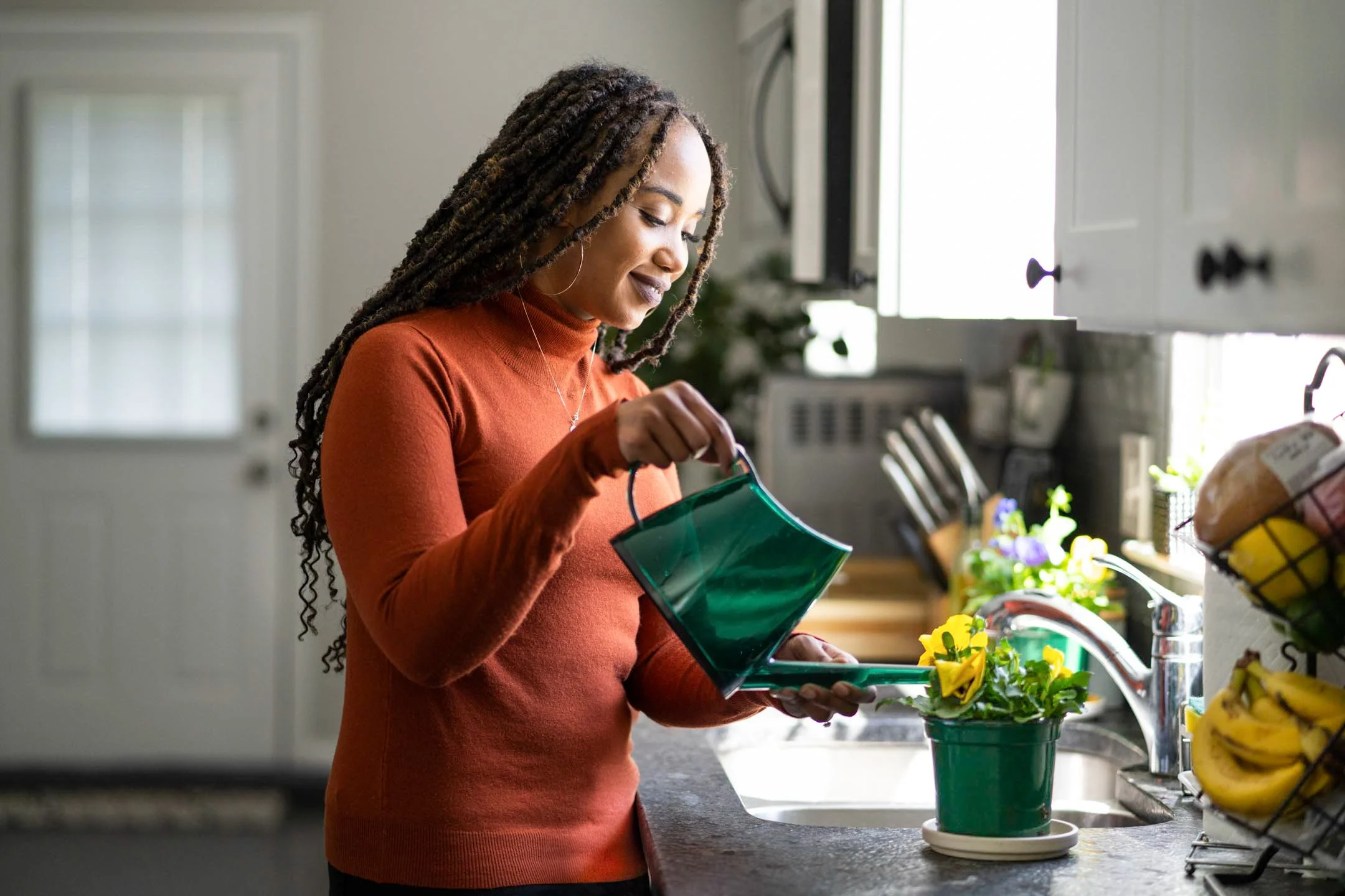 A woman with braided hair watering a potted plant on a kitchen counter.