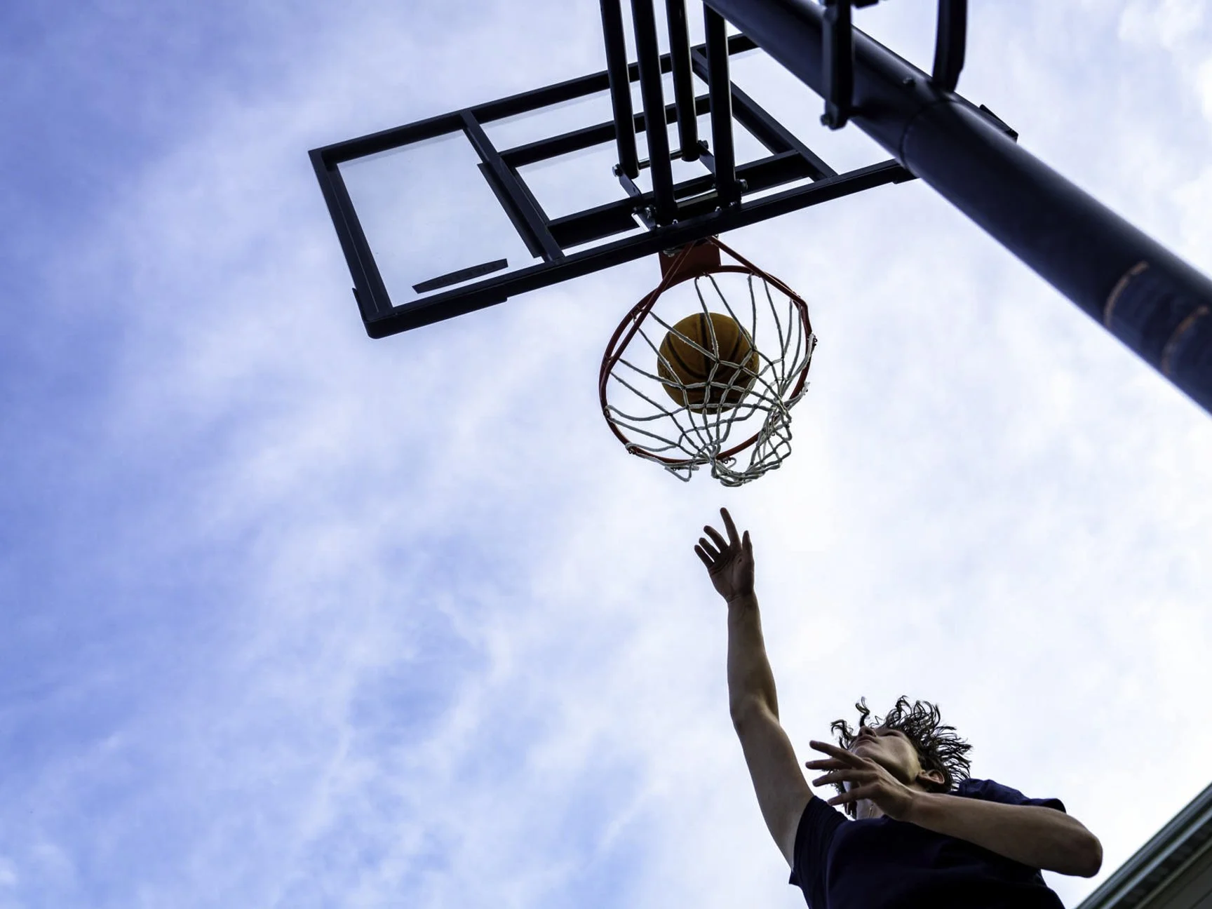 A person playing basketball, shooting a ball into the hoop from below against a partly cloudy sky.