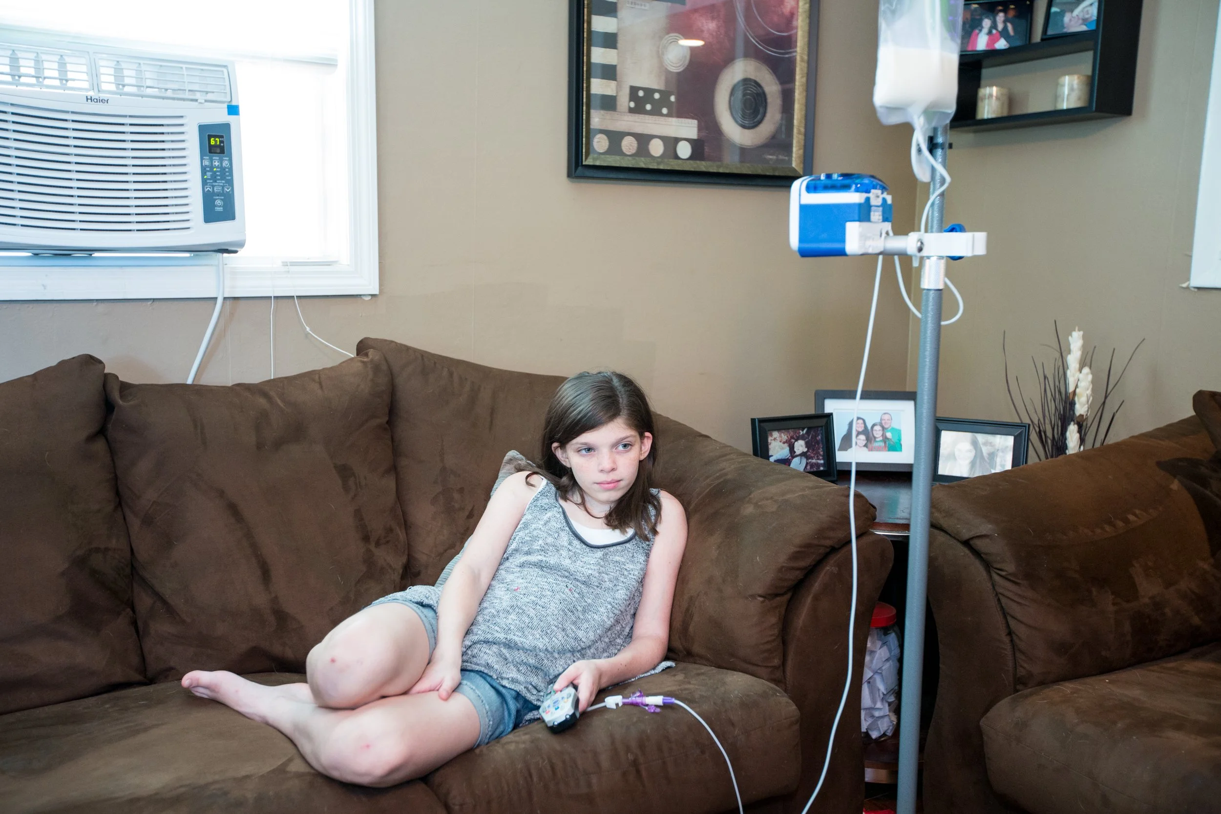 A young girl sitting on a brown couch in a living room, hooked up to IV fluids, with an oxygen unit nearby, watching TV.