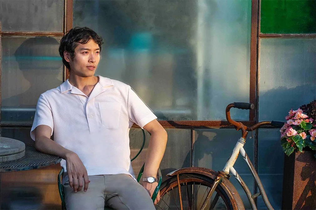 A young man in a white polo shirt sitting outside near a bicycle and a flowerpot with pink flowers, in front of a glass wall.
