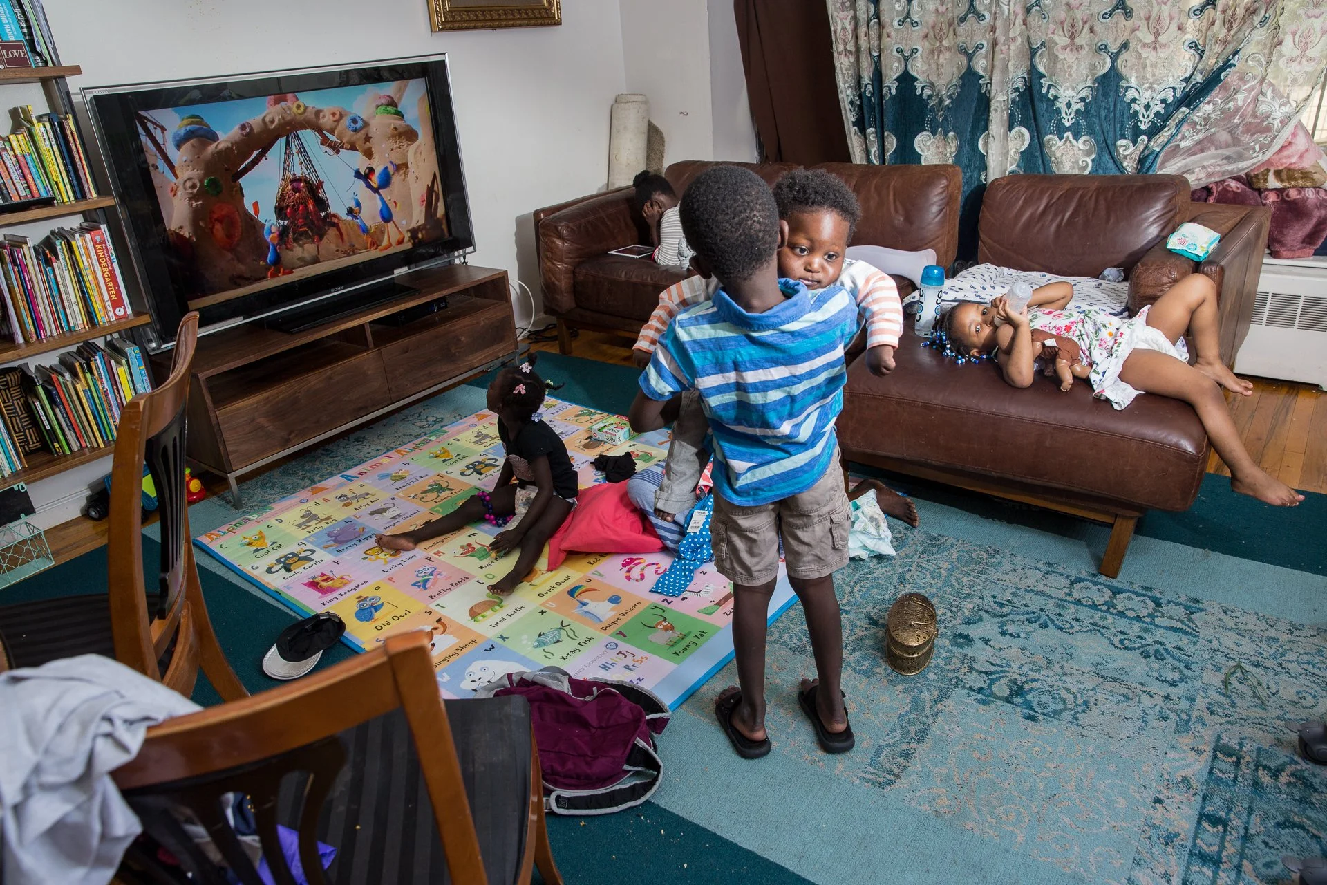 A living room with children playing and relaxing; one child is standing near a couch holding a baby, another is lying on the couch drinking water, and a girl is sitting on a colorful alphabet and number mat.