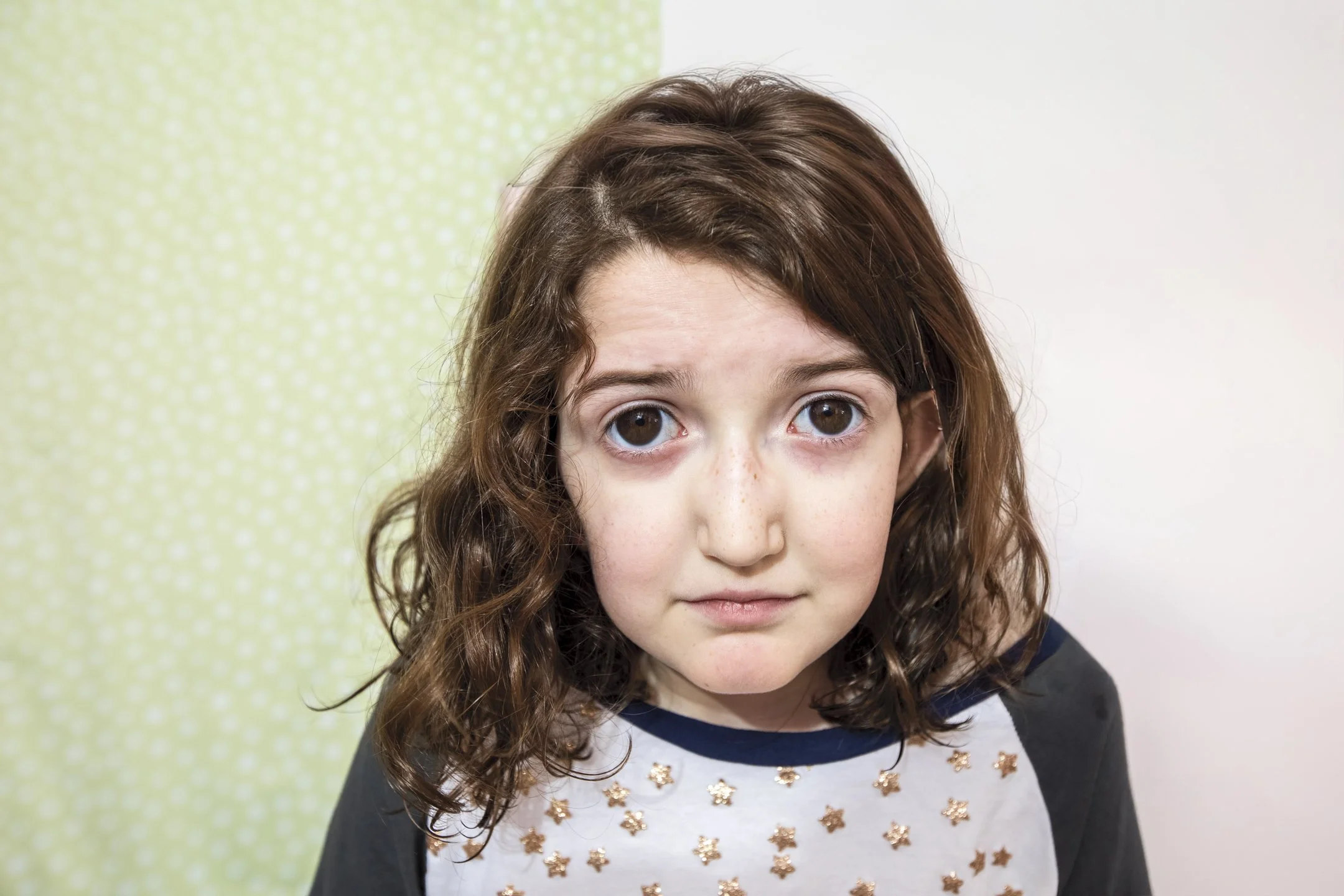 A young girl with curly brown hair and brown eyes, wearing a shirt with small star pattern, looking at the camera with a neutral expression.
