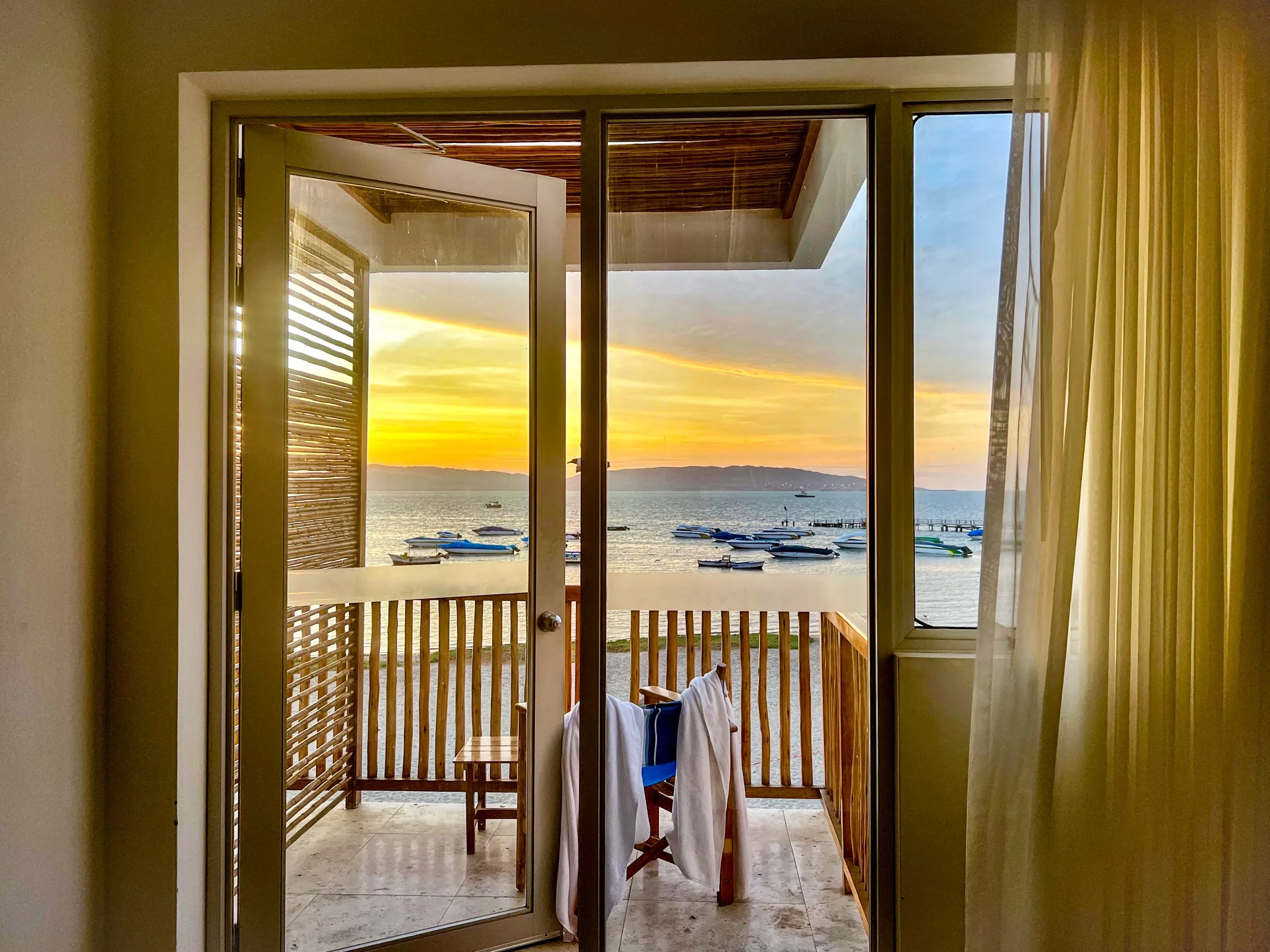 Sunset view through a glass door opening to a balcony with boats on the water and a distant shoreline.