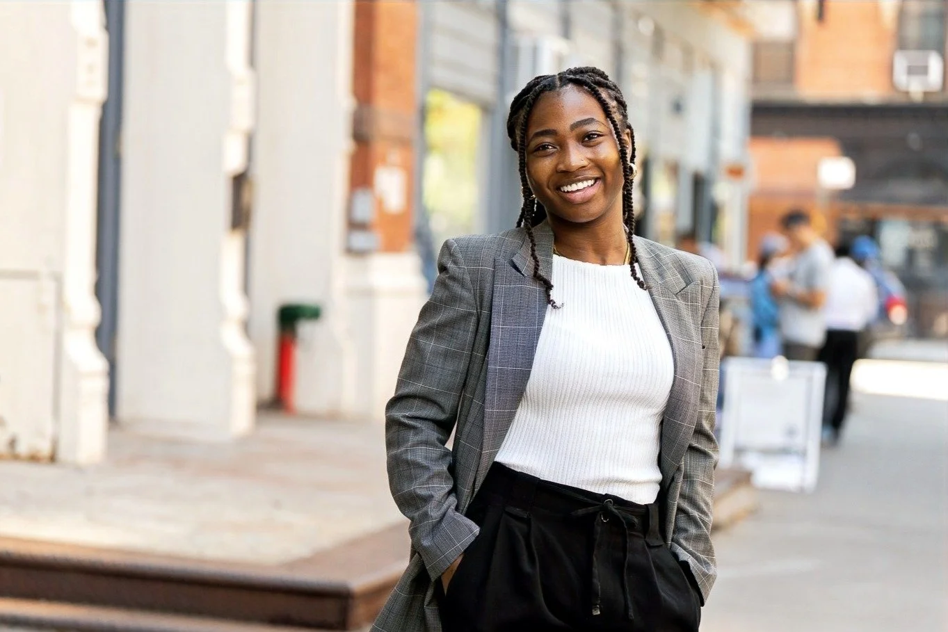 A smiling woman with dreadlocks and dark skin, wearing a gray blazer and white top, standing outdoors on a city street.