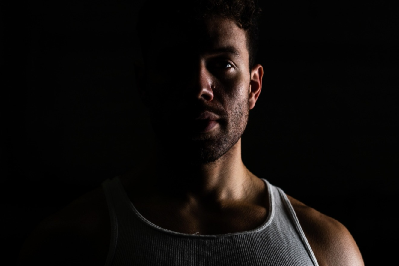 A man with a serious expression, illuminated from the side, wearing a white tank top, against a dark background.