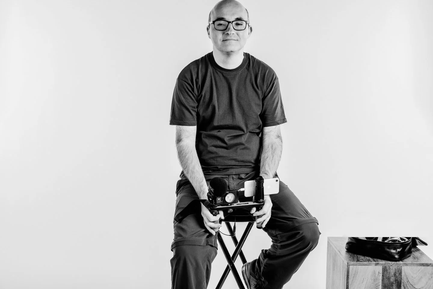 A man with glasses, a black t-shirt, and cargo pants sitting on a stool, holding a gimbal stabilizer with a smartphone attached, in a plain white space.