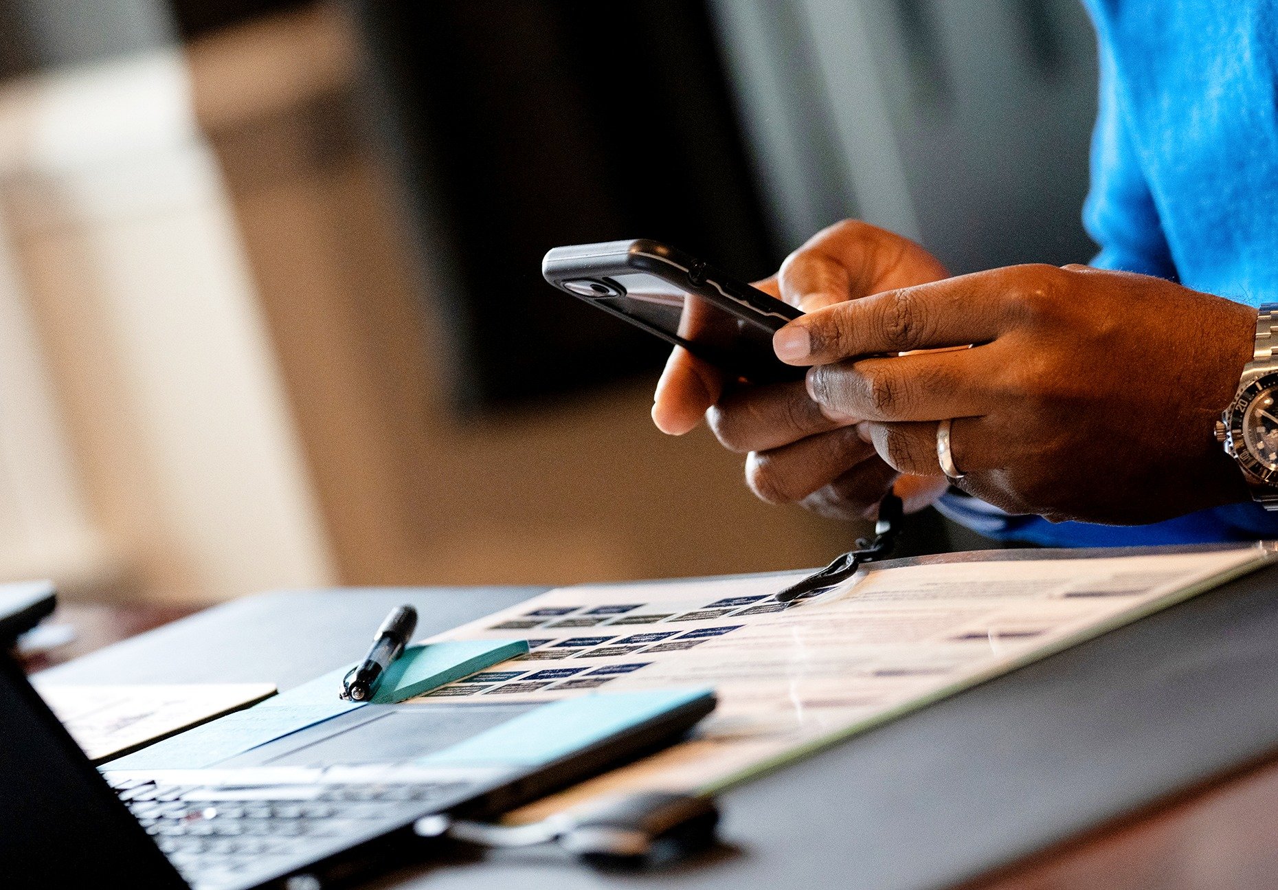 Person using a smartphone with a watch on their wrist, sitting at a desk with a pen, notebook, and printed printed materials.