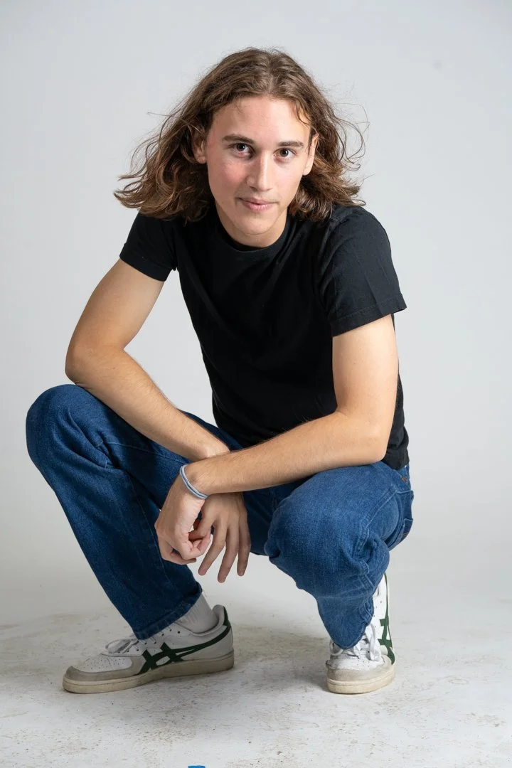 A young person with shoulder-length curly hair, wearing a black T-shirt, blue jeans, and white sneakers, squatting against a plain light gray background.