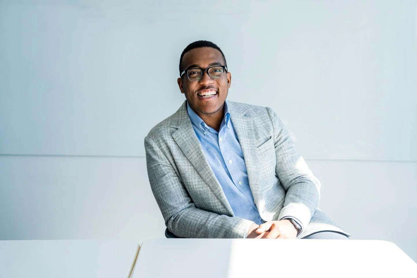 Smiling man in glasses and a gray blazer sitting at a white table in a modern office.