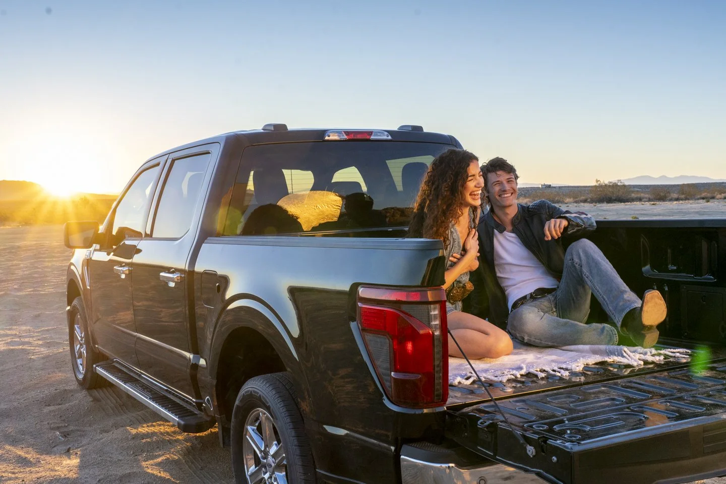 A young couple sitting on the open tailgate of a black pickup truck in a desert at sunset, enjoying each other's company and smiling.