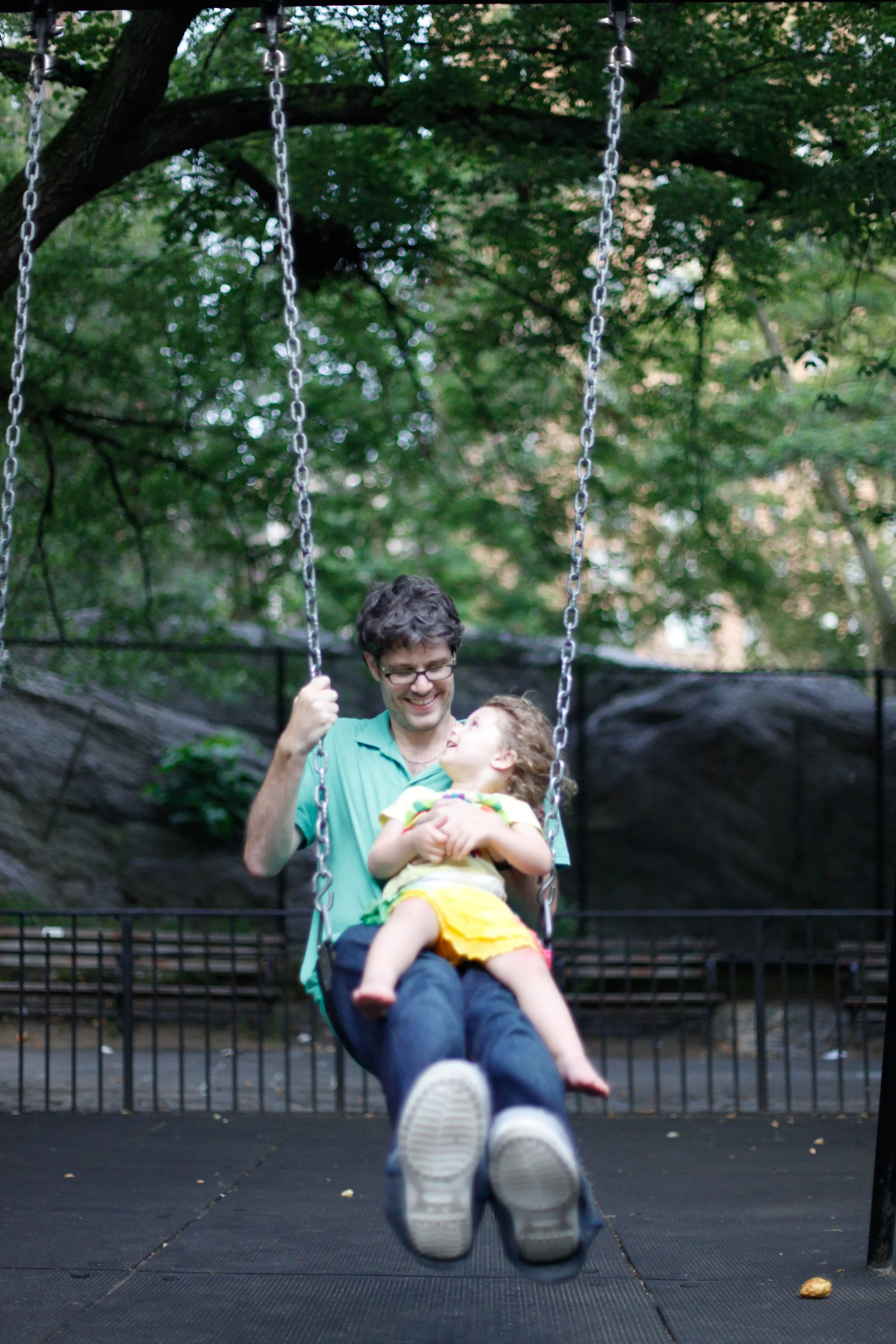 A man and a young girl on a swing, smiling and enjoying themselves in a park with trees and greenery in the background.