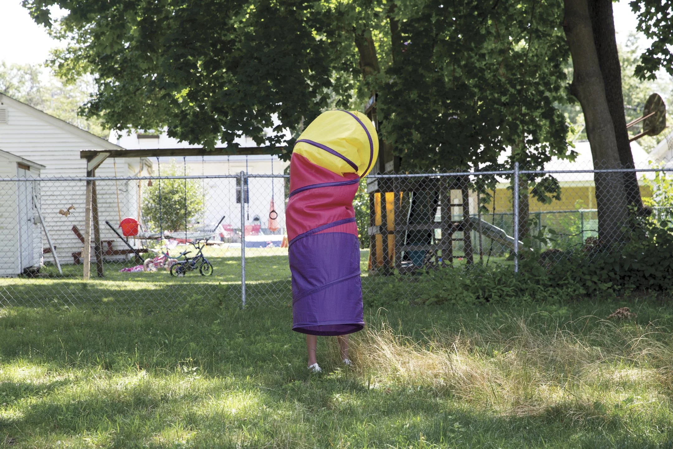Colorful fabric tunnel in a backyard with grass, a chain-link fence, trees, and children's toys.