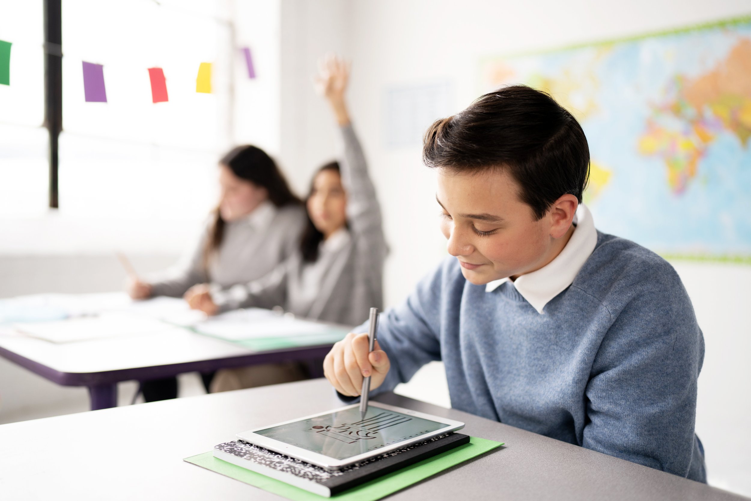 A boy using a tablet in a classroom, with two girls in the background, one raising her hand.