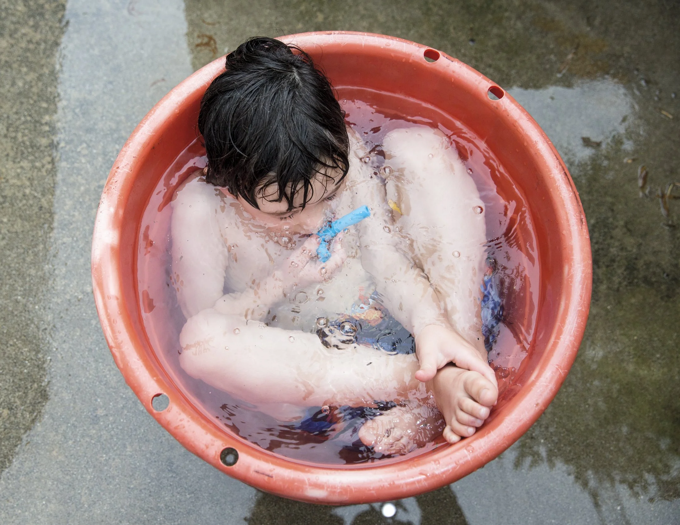 A young boy with wet dark hair is sitting in a round orange plastic tub filled with water, playing underwater with a blue toy. The tub is placed on a concrete surface outdoors.