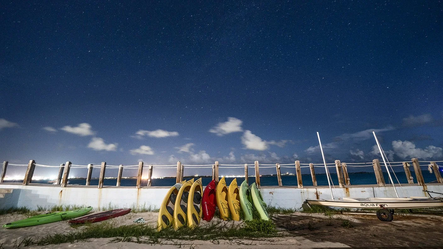 Kayaks and a small sailboat on a sandy beach at night, with a wooden fence and a starry sky in the background.