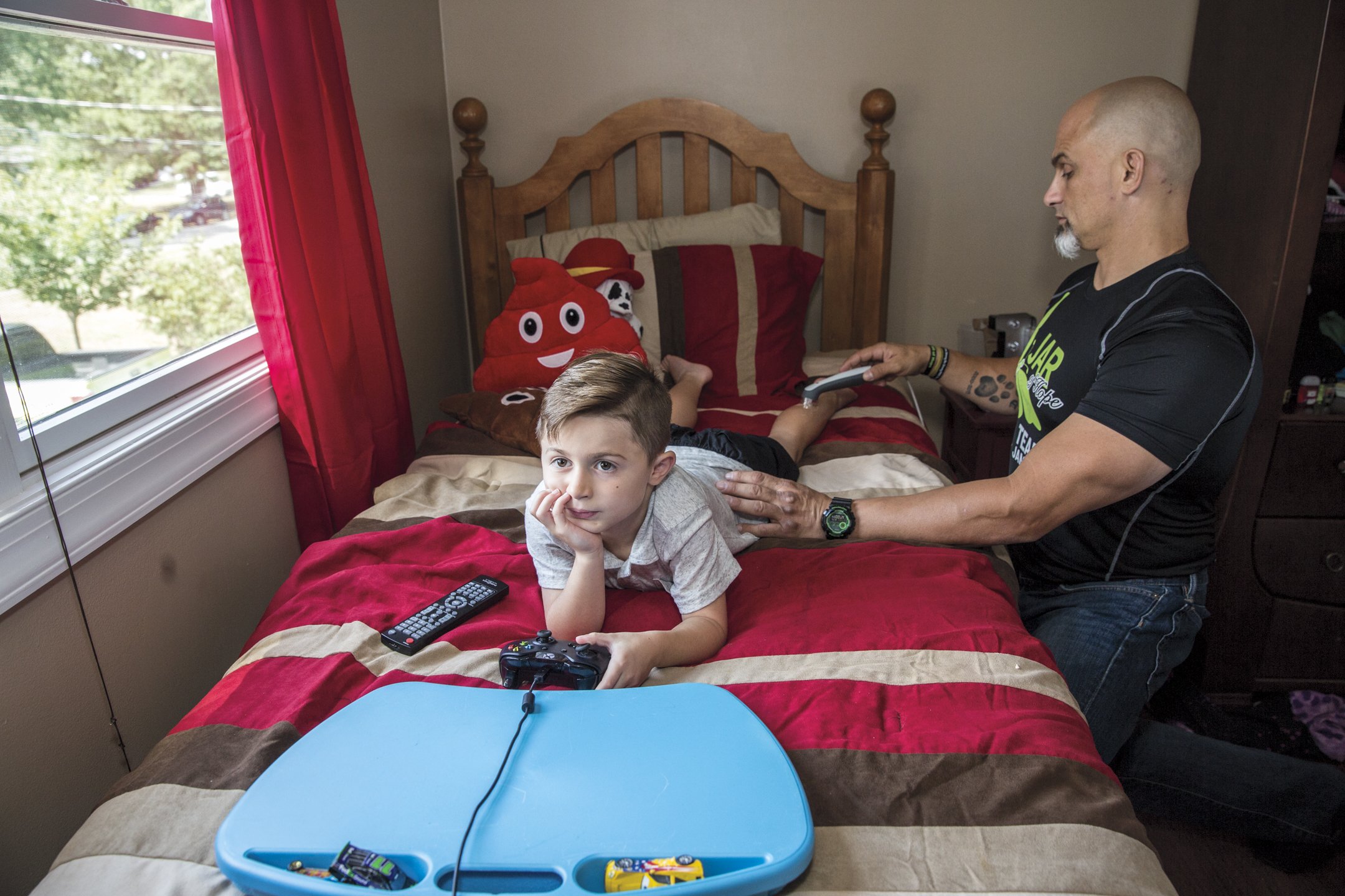 A young boy lying on a bed with a thoughtful expression, holding a gaming controller. An adult man, possibly his father, is vacuuming the bed. The bed has red, tan, and brown bedding, and there are plush toys, including a poop emoji pillow, on the he