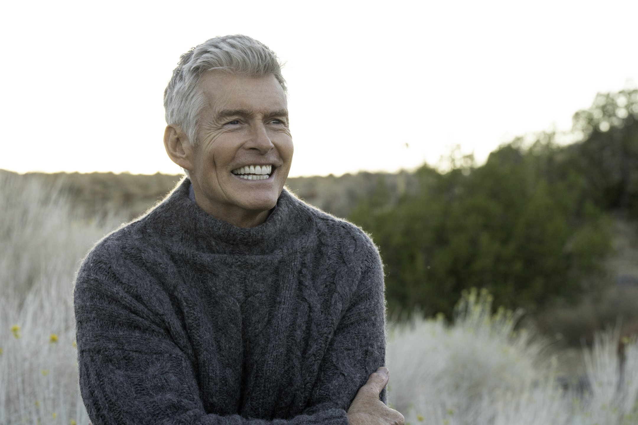 An elderly man with gray hair and a wide smile wearing a dark gray knitted sweater outdoors in a natural landscape.