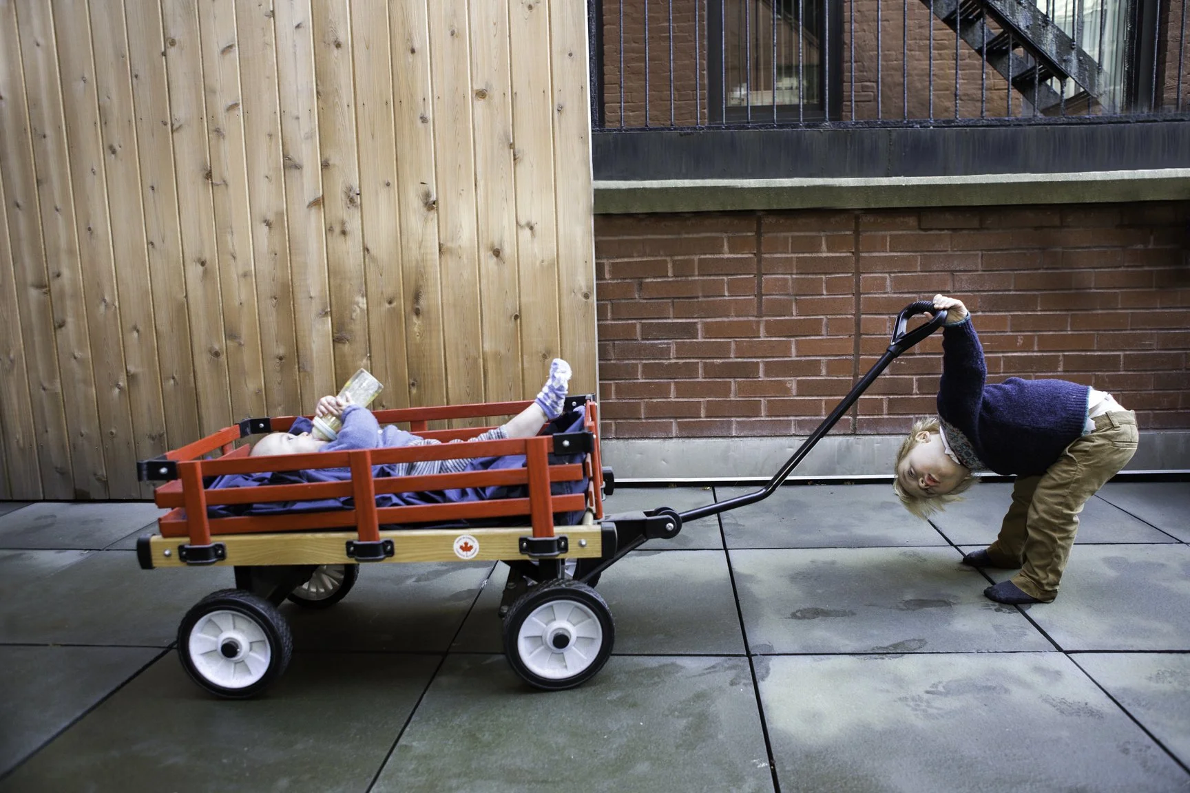 A young boy pulling a red wagon with a younger child lying inside reading a book.