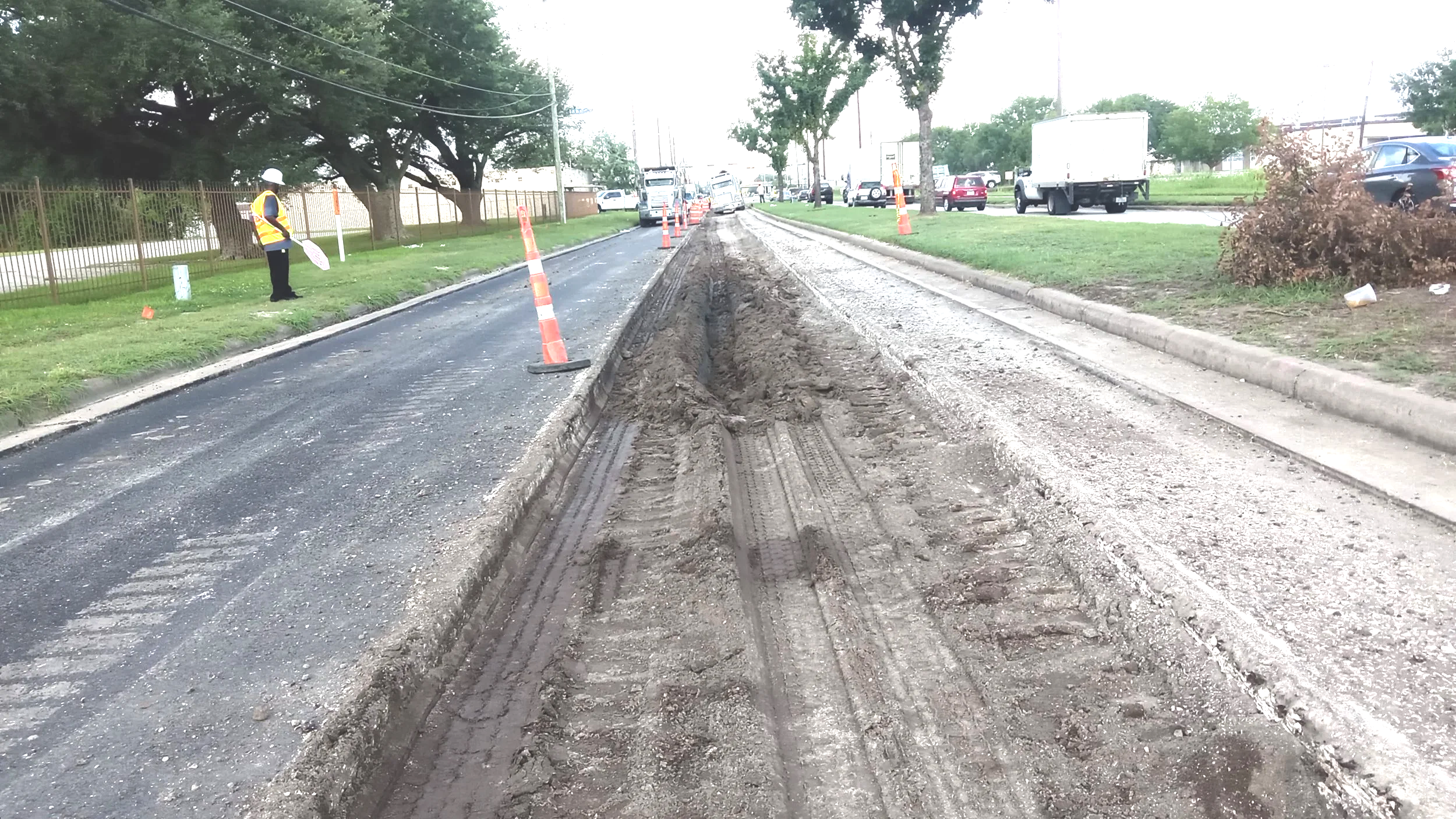 Construction site on a city street with a dug-up middle lane, orange cones, a worker wearing a hard hat and safety vest holding a sign, cars on the road, trees, and a fence on the left side.