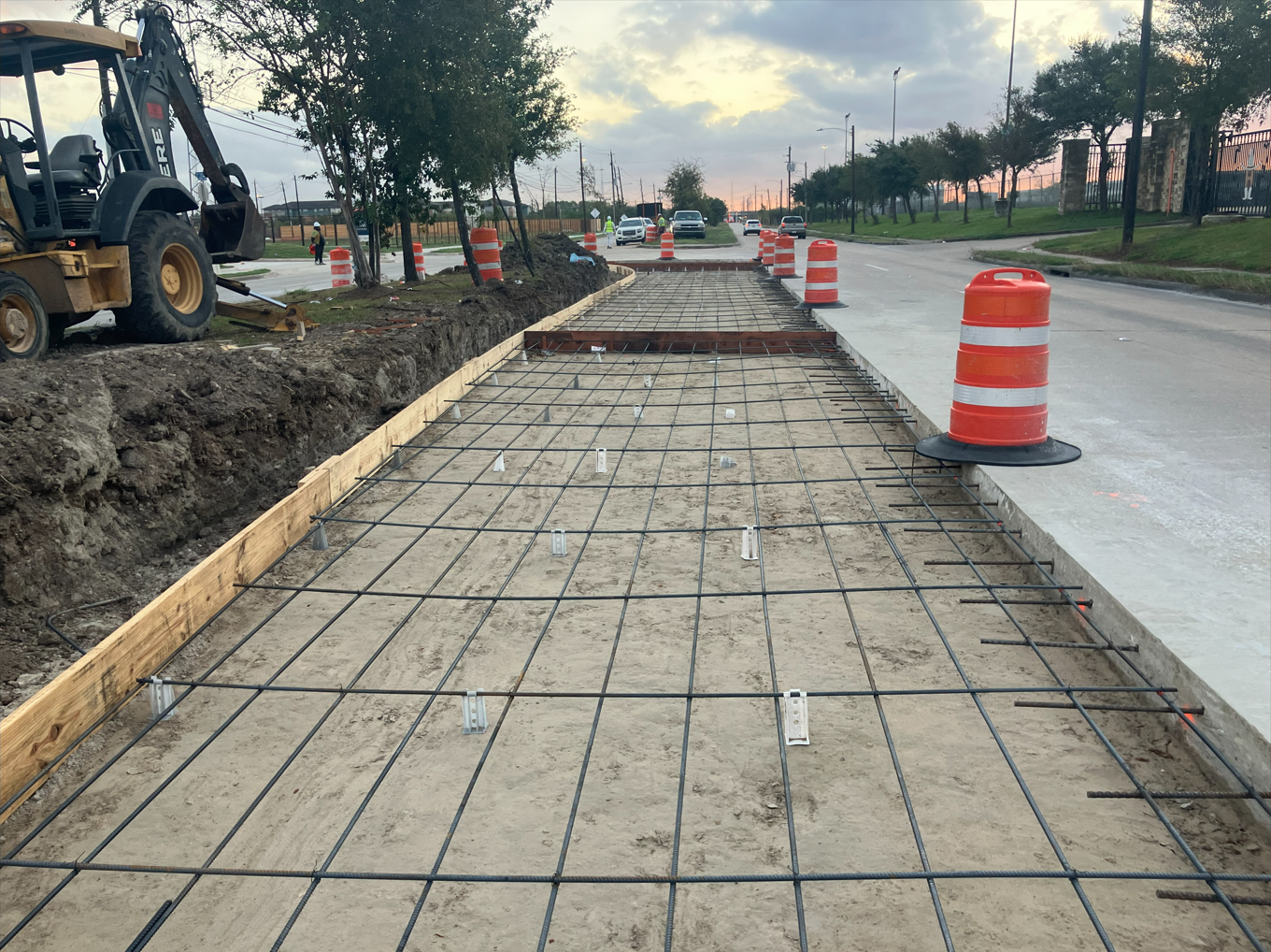 Road construction site with rebar framework for concrete sidewalk, orange traffic barrels, construction machine, trees, and cars on the street during sunset.