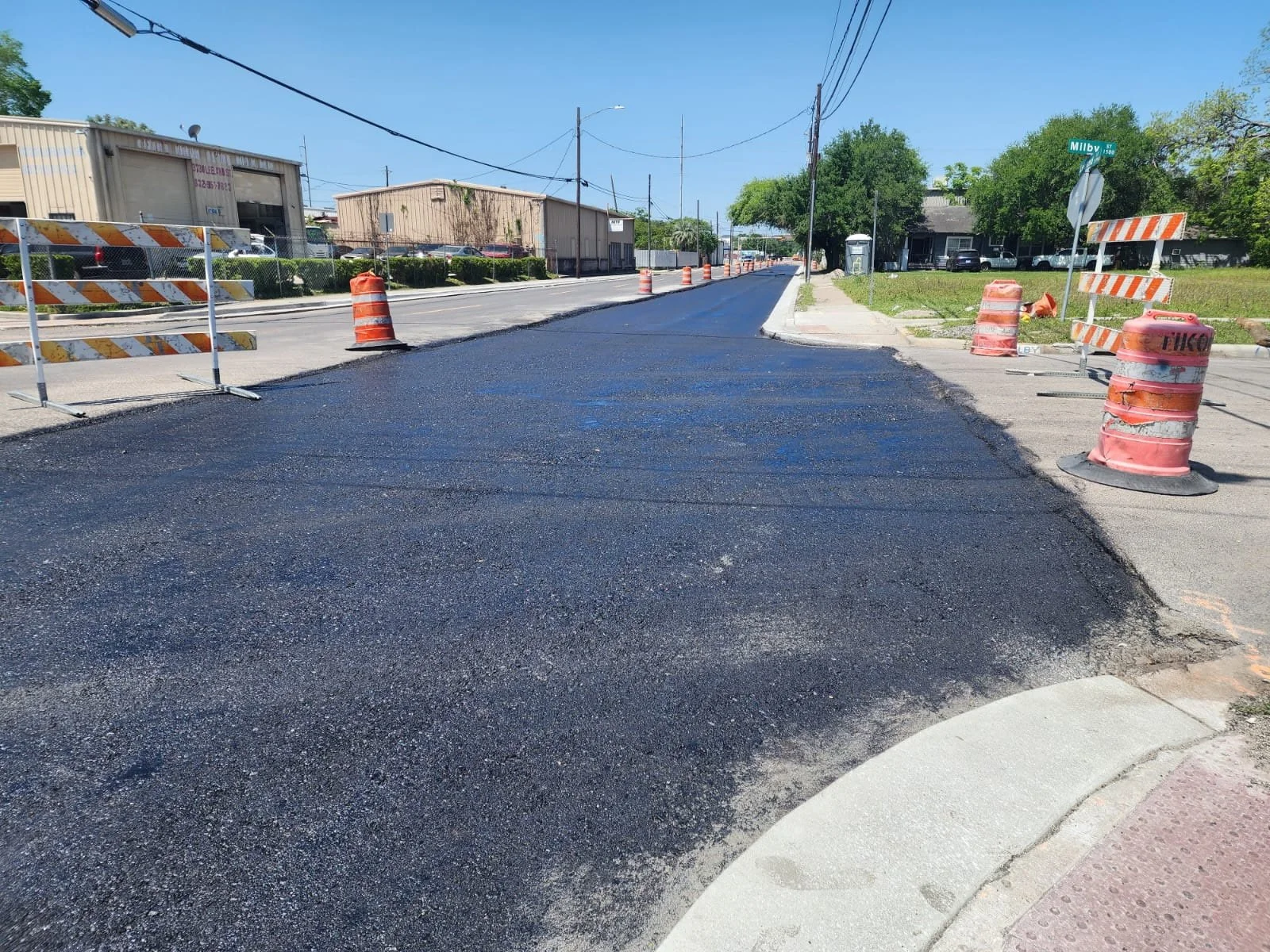A street undergoing road construction, with fresh black asphalt, orange barrels, and barricades, in a residential and commercial area with trees and buildings.