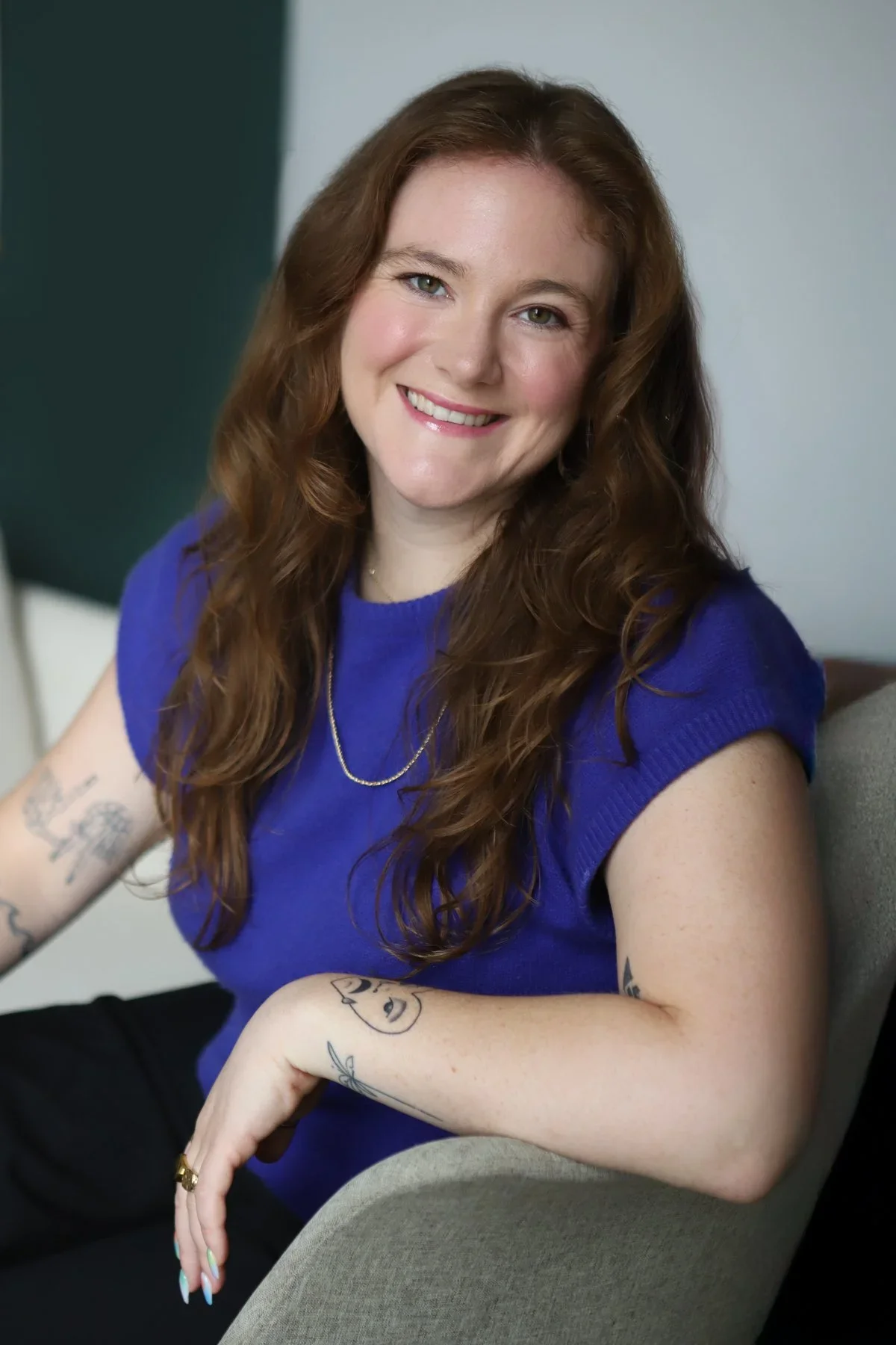 A woman with long red hair wearing a blue sleeveless top, smiling, sitting on a gray chair, with visible tattoos on her arms and a gold necklace.