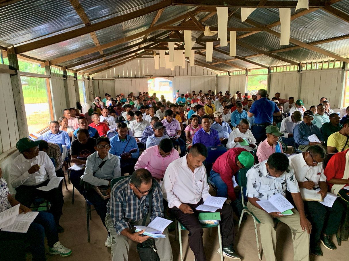 A large group of people attending a meeting or conference in a spacious barn-like room, seated in rows, some reading documents, with a person standing and speaking at the front.