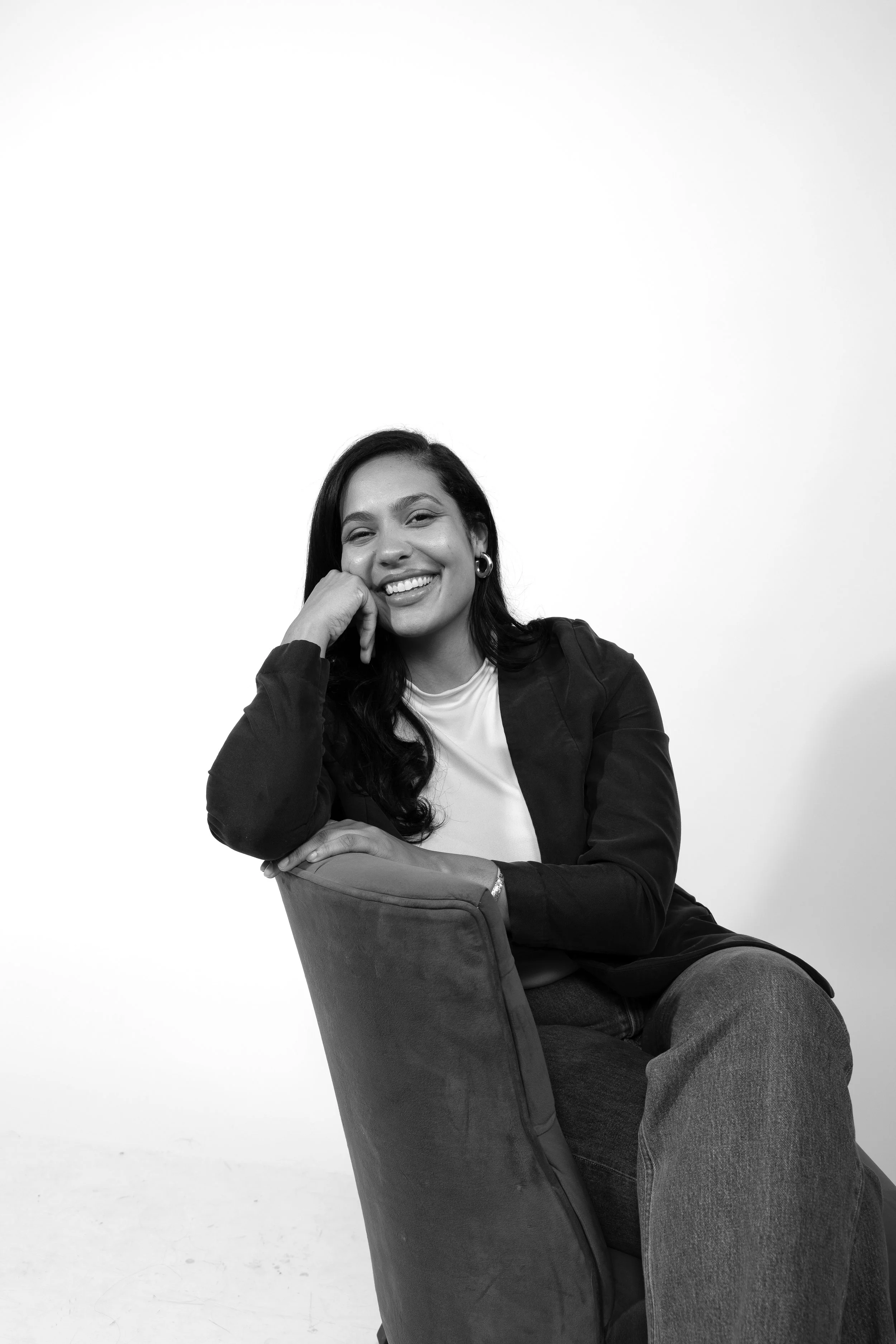 Portrait of a smiling woman with dark hair, wearing earrings, sitting in a chair with her chin resting on her thumb against a plain white background.
