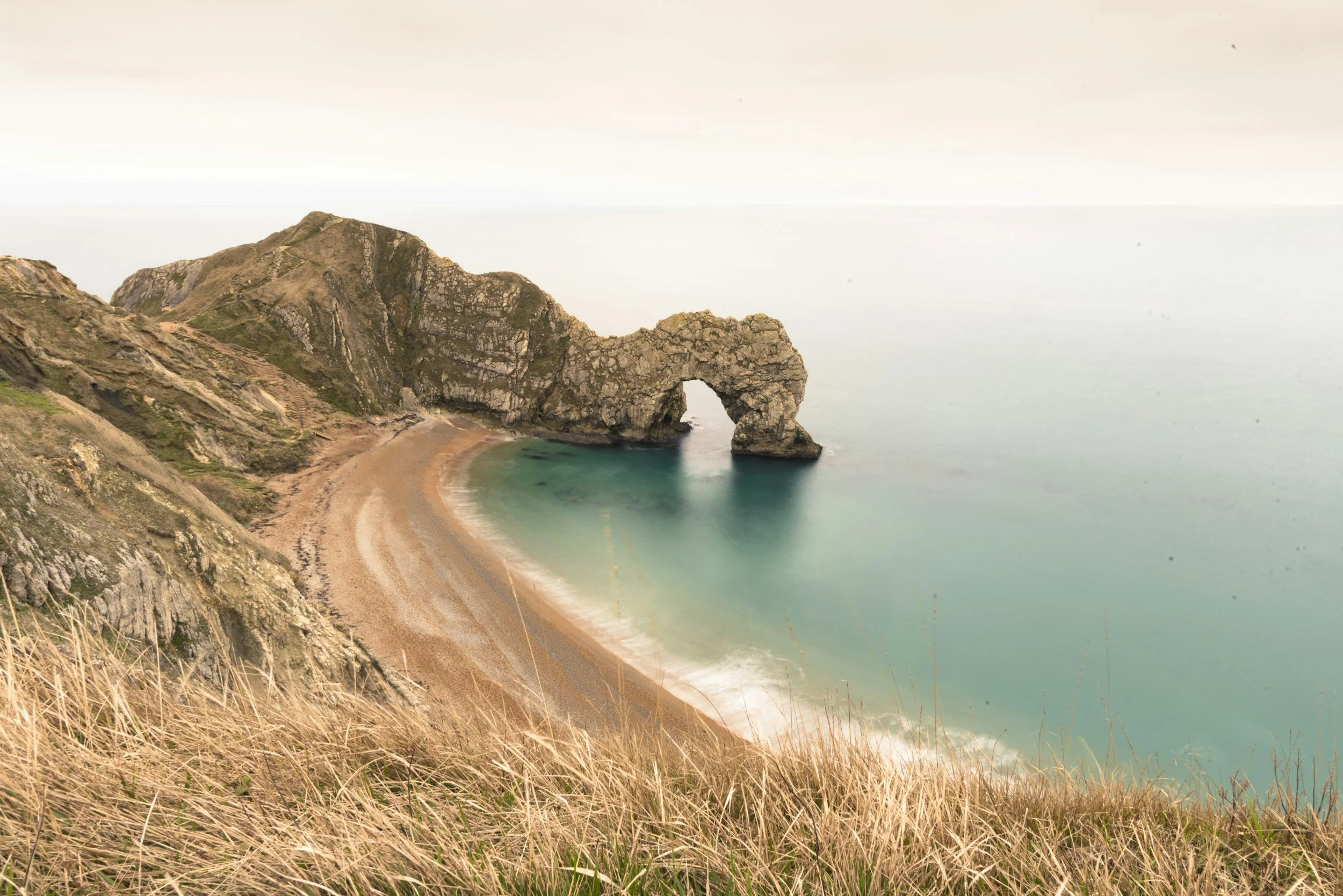 Cliffs and rocky arch on a beach with a sandy shoreline and turquoise ocean water, seen from a grassy hill.