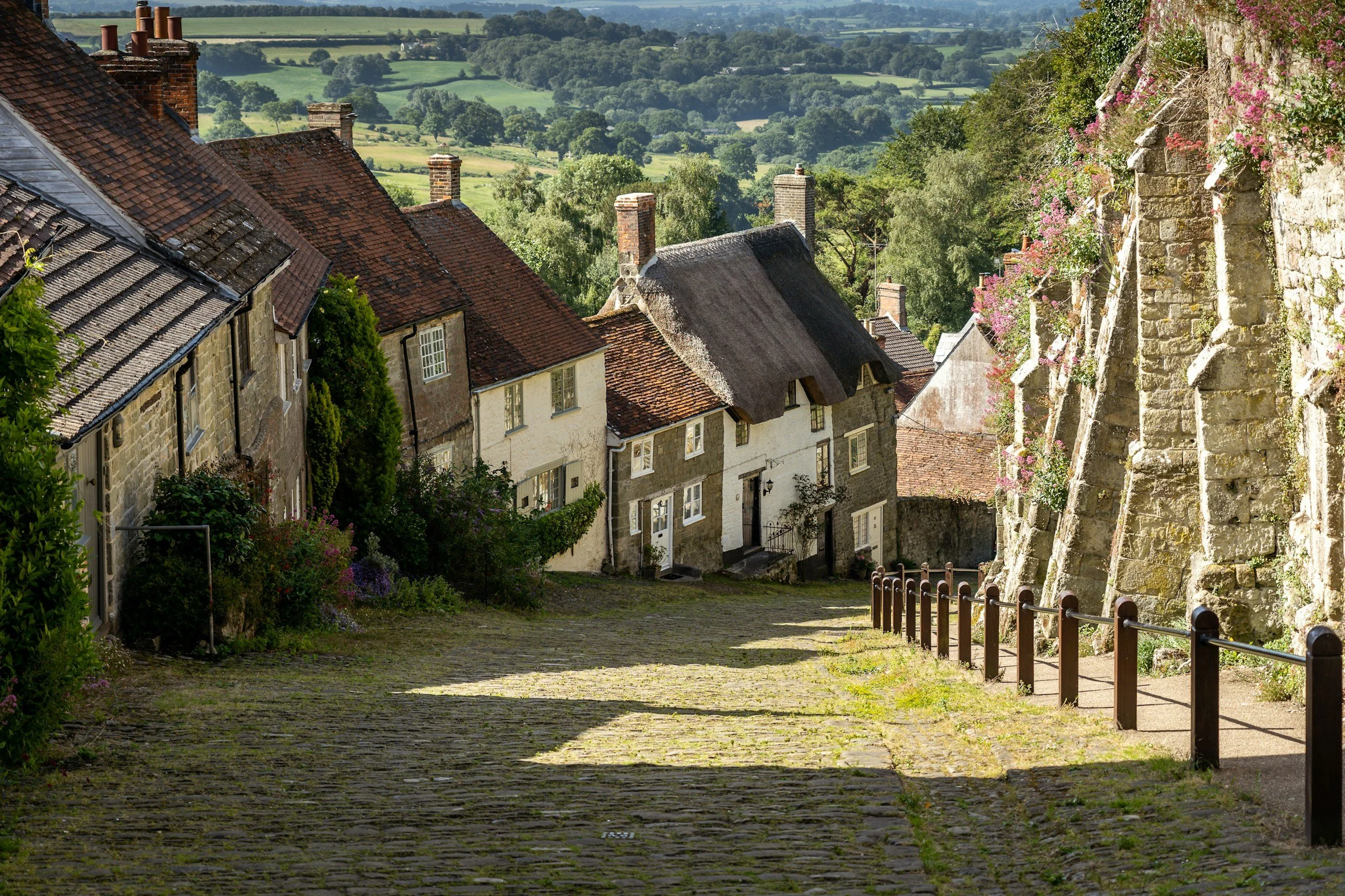 A cobblestone street slopes downward next to historic houses with pitched roofs, some overhanging, and a stone wall covered in flowering plants. In the background are lush green fields and rolling hills under a clear sky.