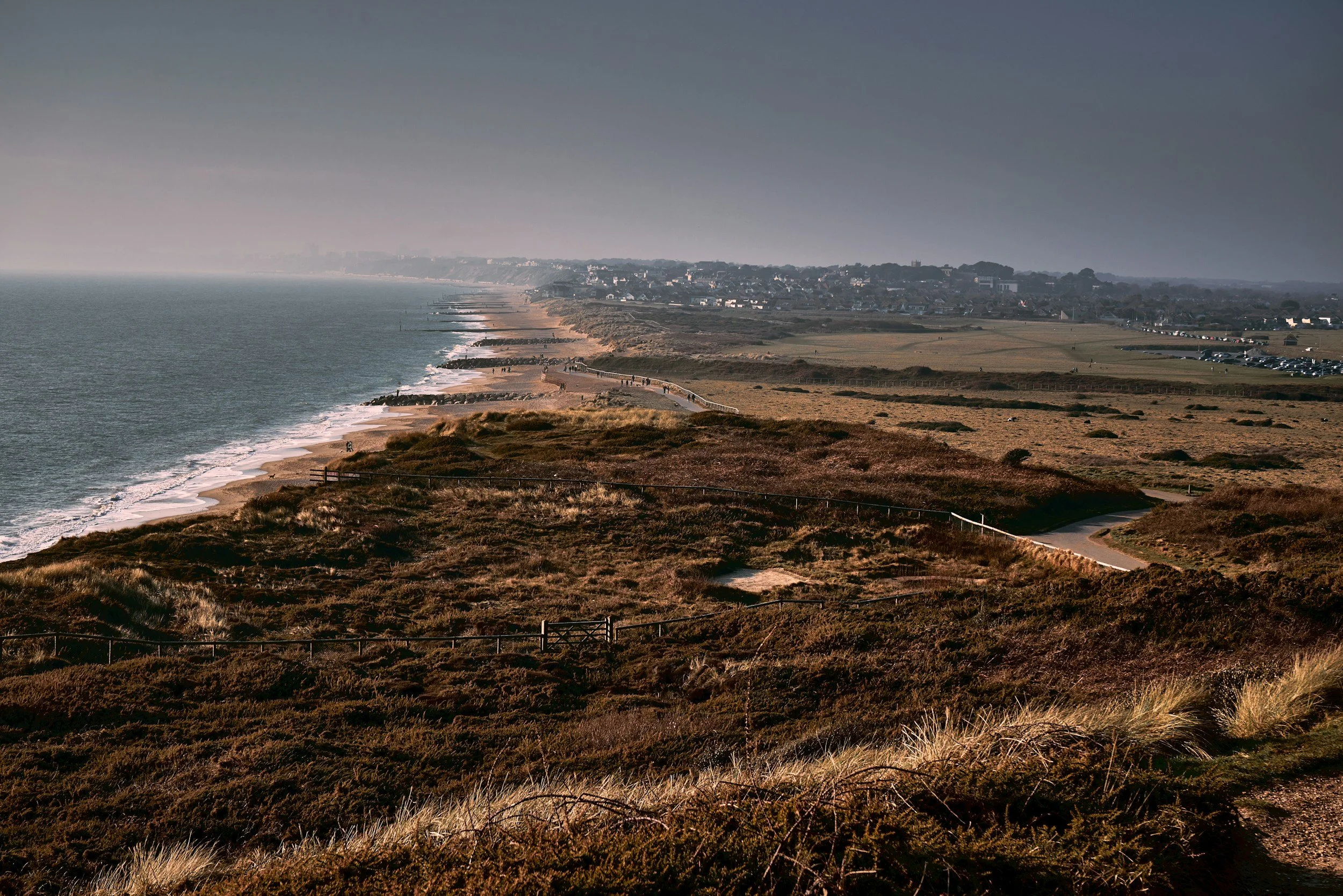 A coastal landscape with a sandy beach, waves, grassy dunes, and a small town in the distance under a cloudy sky.