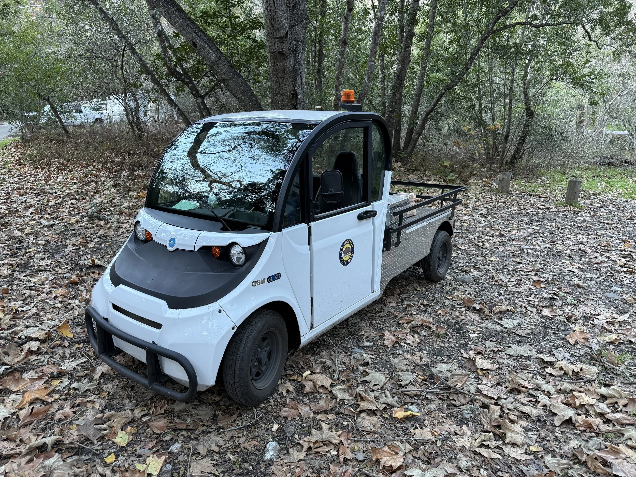 A small electric utility vehicle with a white body and black accents parked on a leaf-covered ground in a wooded area. The vehicle has a cargo bed at the back and a sticker on the door revealing it belongs to the California State Parks