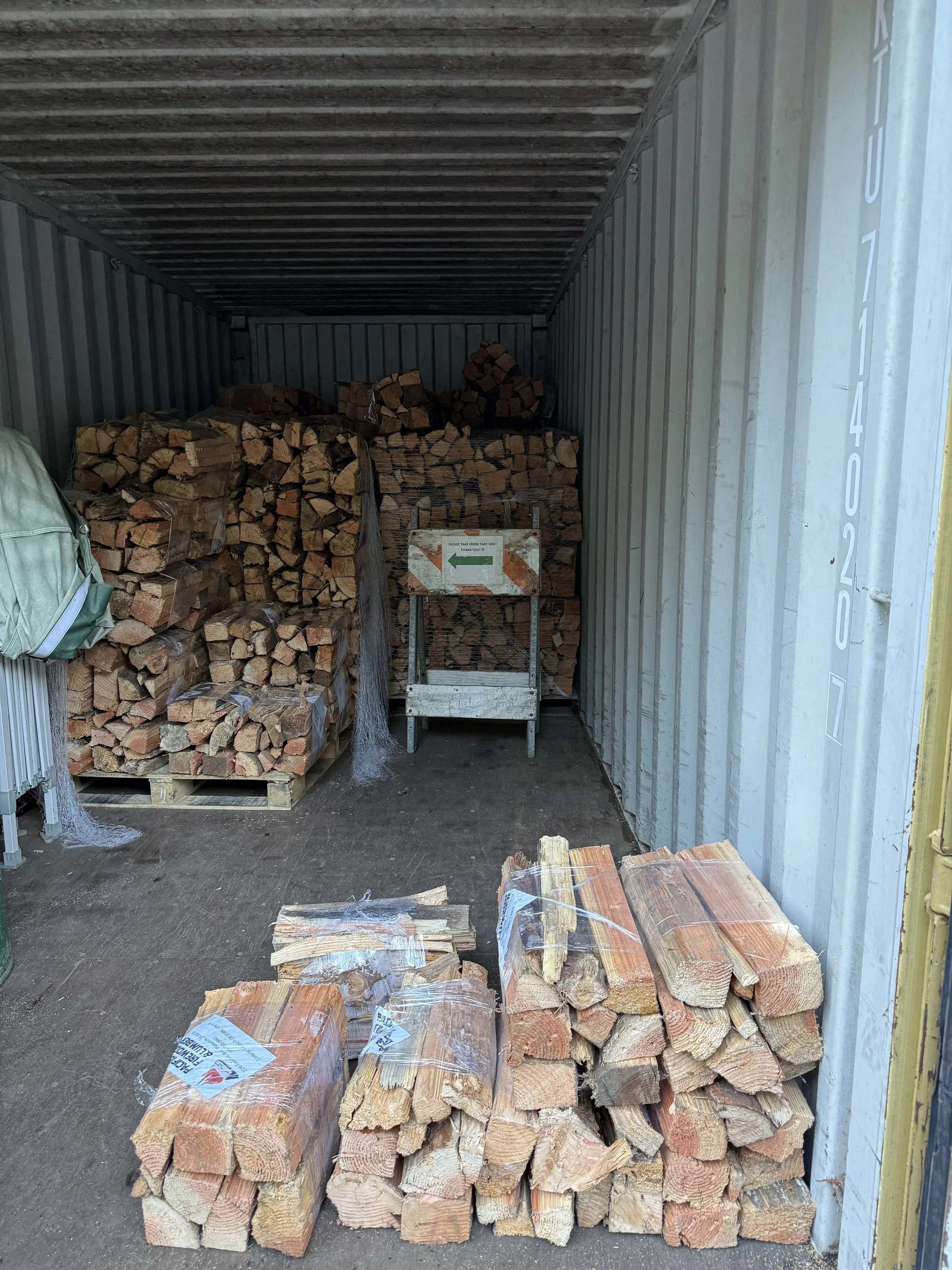 Stacks of cut firewood inside a shipping container with some wood on the ground and a small sign on a cart at the back.