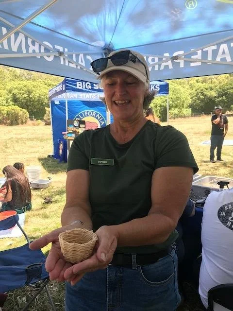 A smiling woman wearing a beige cap and sunglasses on her head, holding a small woven basket, outdoors at a park or festival with tents in the background.