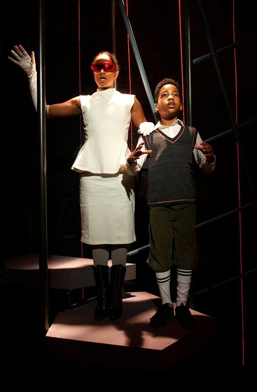 Two children, a girl and a boy, standing on a platform in a dark space with red vertical light strips; the girl is wearing a white dress, glasses, gloves, and black boots, while the boy is dressed in a school uniform with a vest, shirt, shorts, knee-high socks, and black shoes.