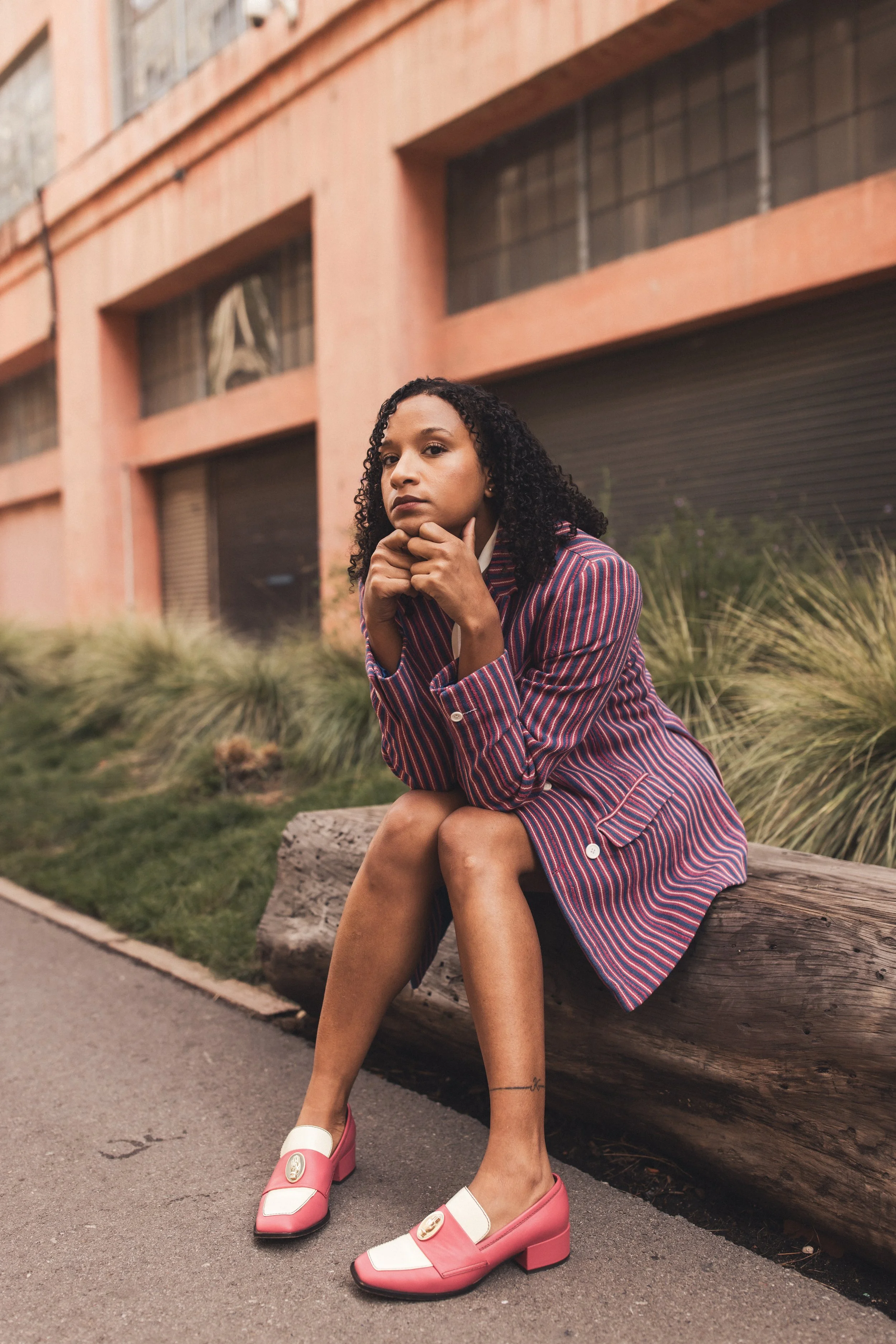 A woman with curly black hair sitting on a log in an urban outdoor setting, wearing a striped blazer and pink and white shoes.