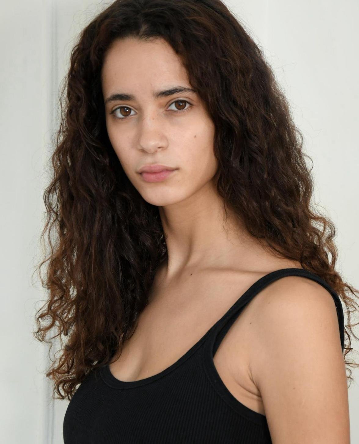 A young woman with long, curly dark hair wearing a black tank top, facing the camera with a neutral expression.