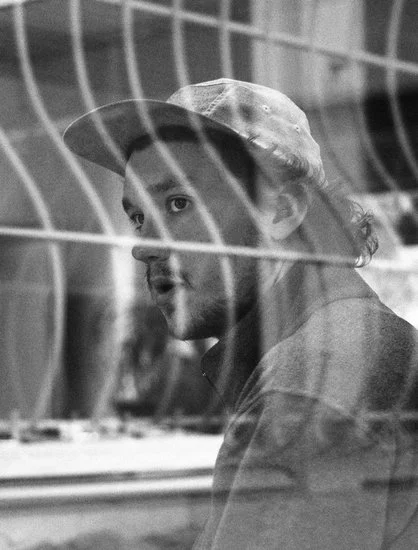 A man with a beard and a hat looking through a wire fence.
