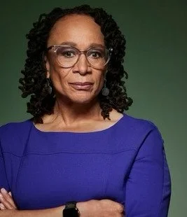 Portrait of an African American woman with curly hair wearing glasses and a blue top, against a green background.
