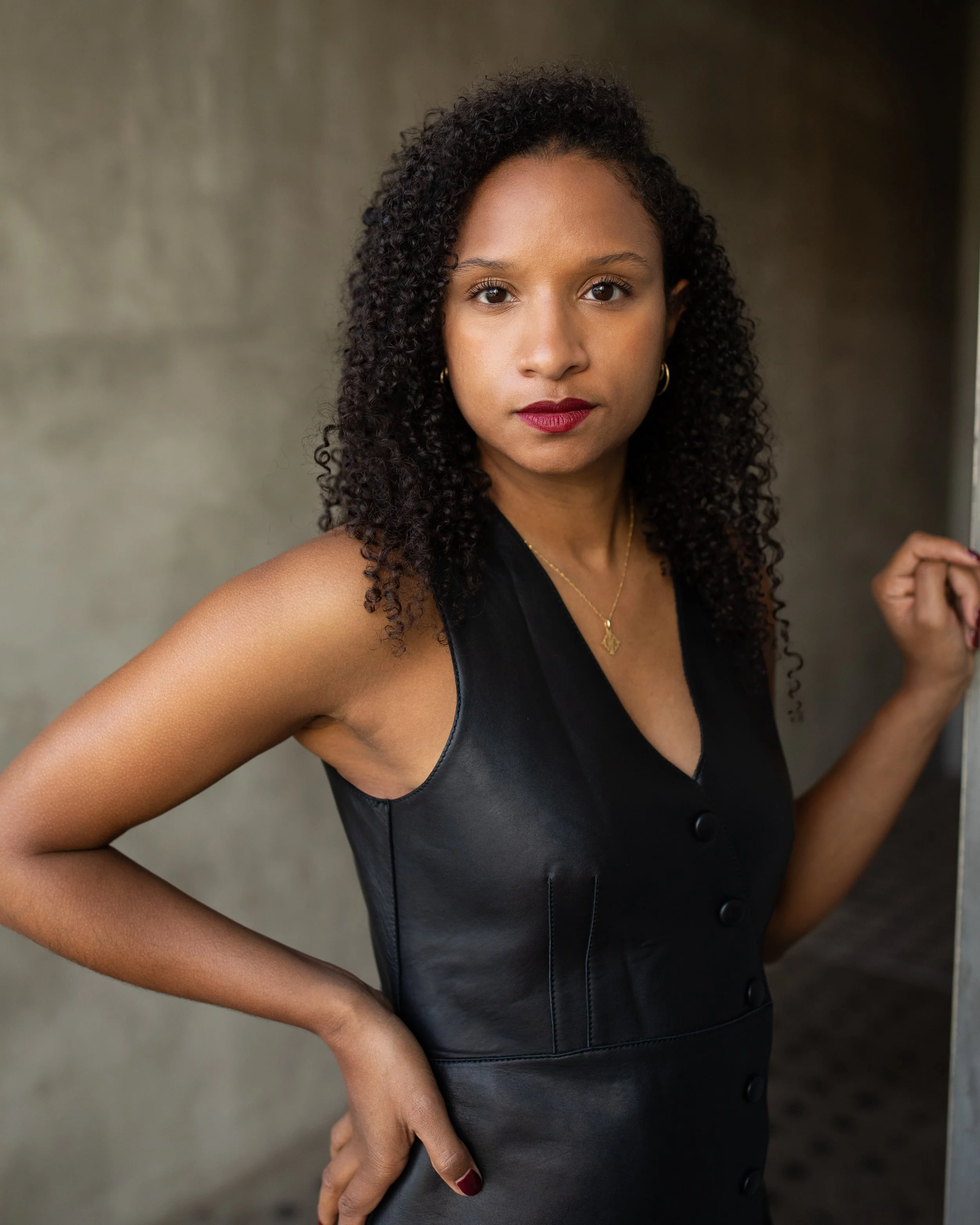 A woman with curly hair and dark skin looking directly at the camera, wearing a black leather sleeveless dress, standing beside a wall.