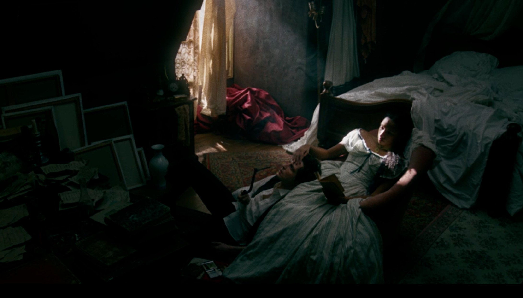 Jeanne Duval and Charles Baudelaire relax on a bed in a dimly lit room, sunlight coming through a window illuminating part of the space with picture frames on a side table.