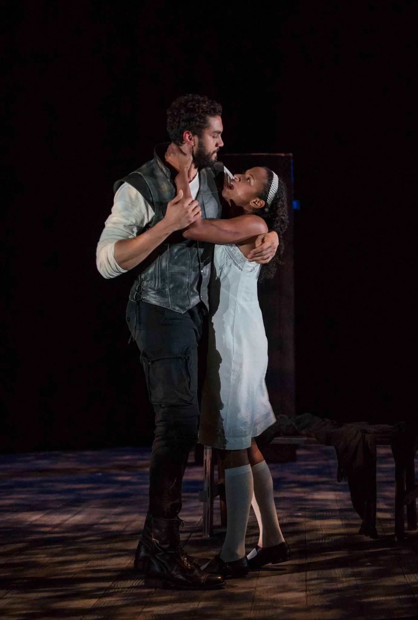 A man and a young girl are performing together on stage, with the girl reaching up to touch the man's face. The man is dressed in dark pants, boots, a white shirt, and a black leather vest, and the girl is wearing a white dress and socks. The scene appears emotional and theatrical.