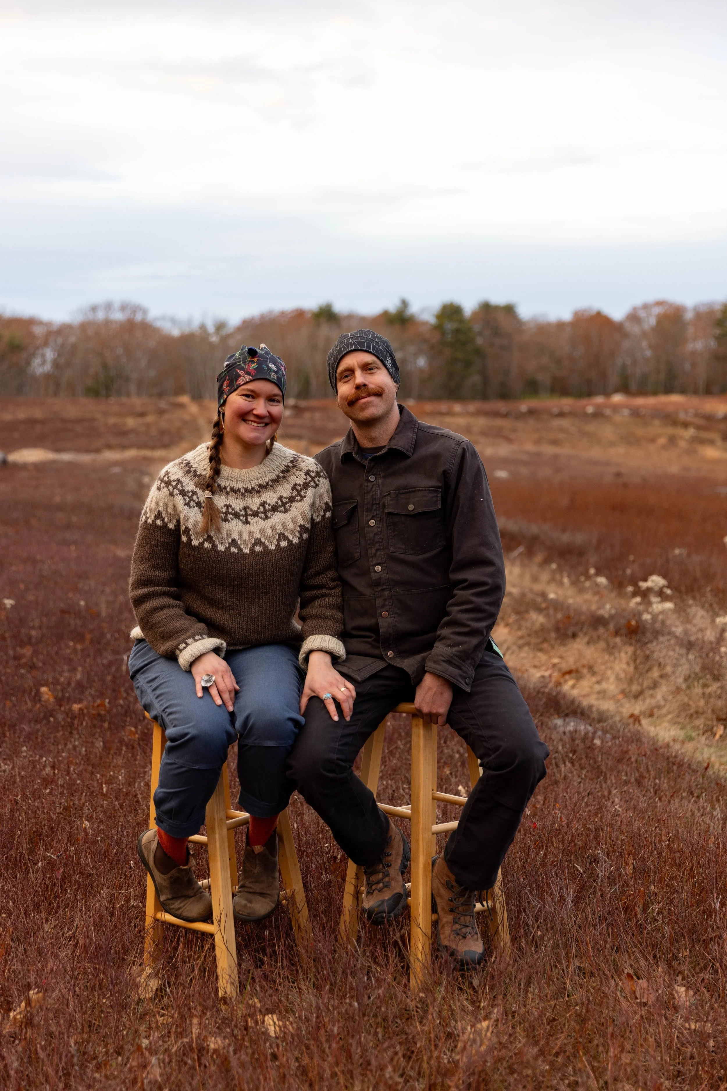 A man and a woman sitting on wooden stools outdoors with a autumn landscape and cloudy sky in background.