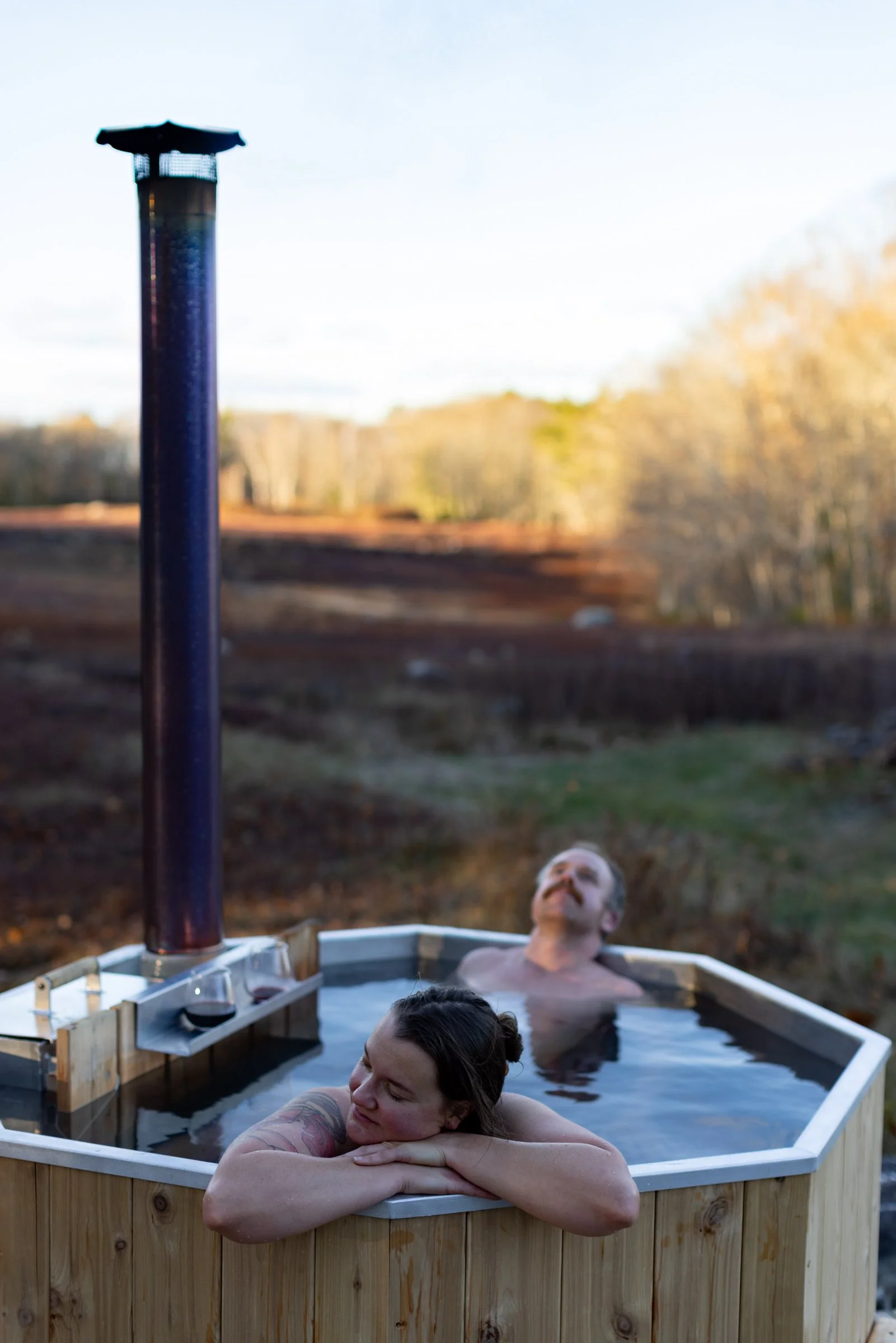 Two people relaxing in an outdoor wooden hot tub, one woman resting her head on the edge and a man floating in the water, with a scenic landscape and trees in the background.