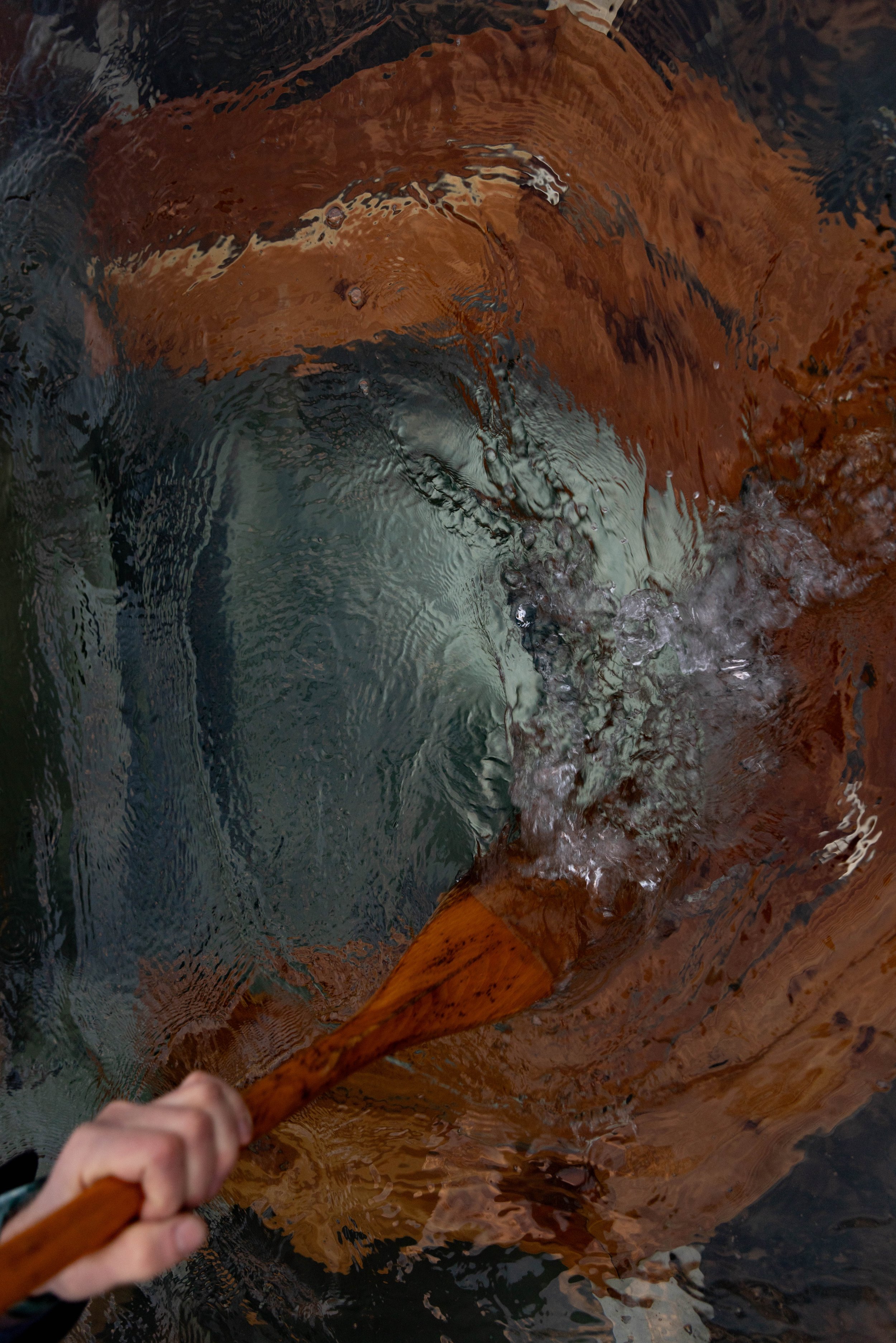 A person using a wooden paddle to stir water in a wood fired hot tub, creating ripples on the surface.