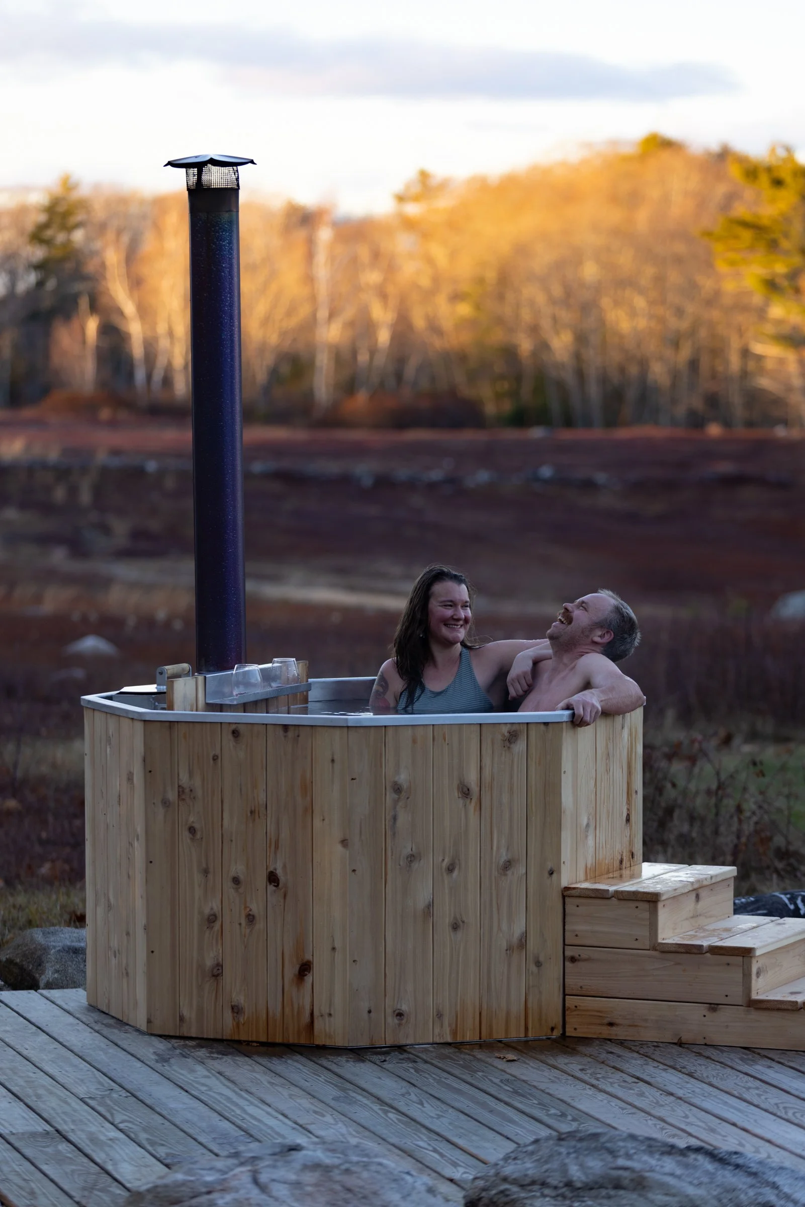 A woman and a man enjoying a wood fired hot tub outdoors in a natural setting with trees and rolling hills in the background.