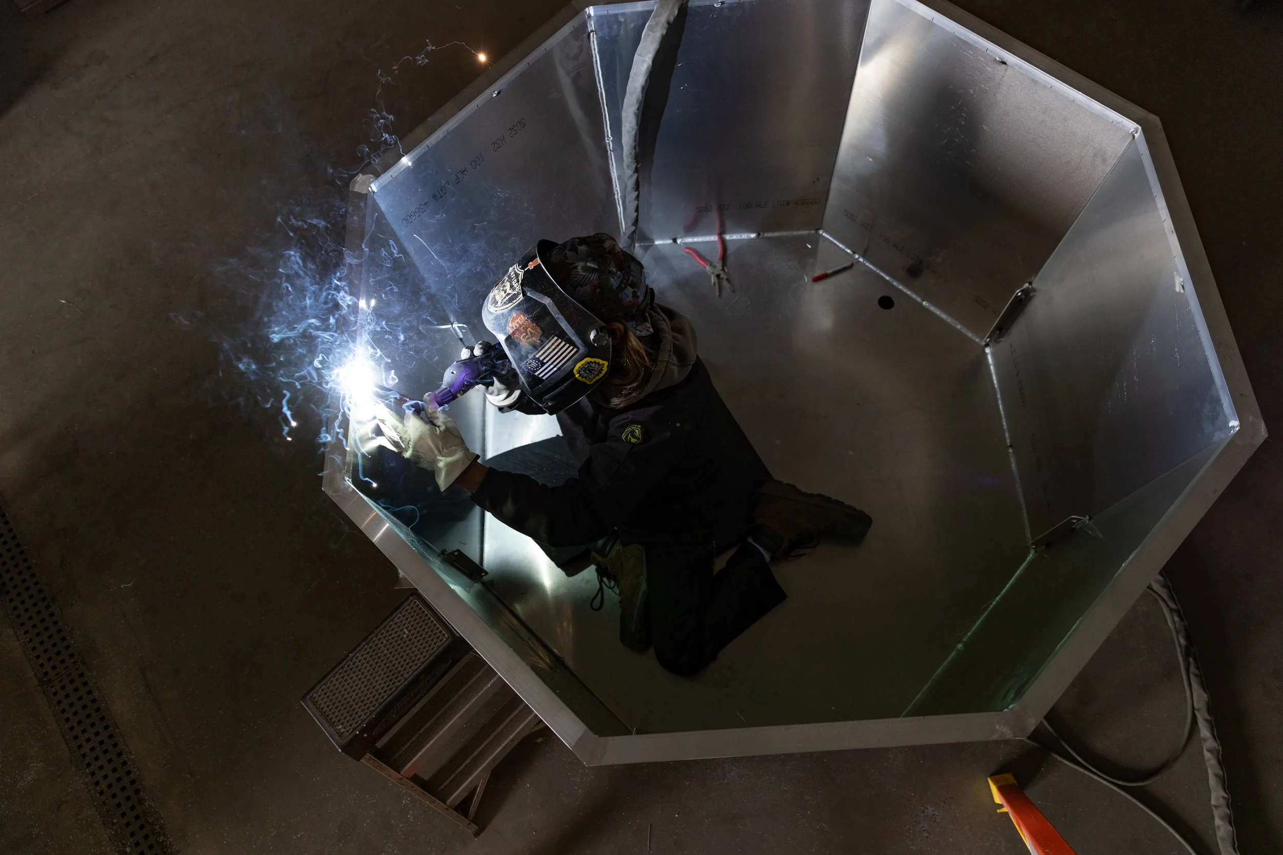 A person welding inside a large, metallic, octagonal enclosure, wearing protective gear including a welding helmet and gloves, with sparks and bright light from the welding process.
