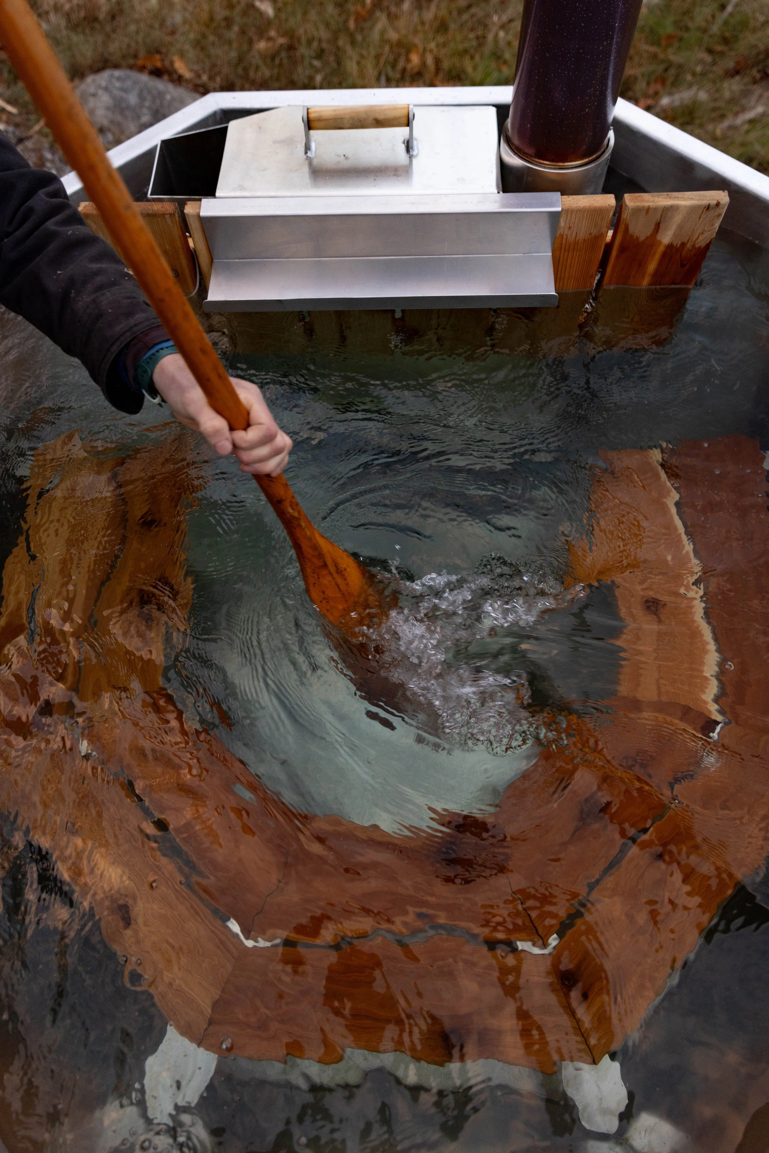 A person stirring water with a wooden paddle in an outdoor wood fired hot tub.