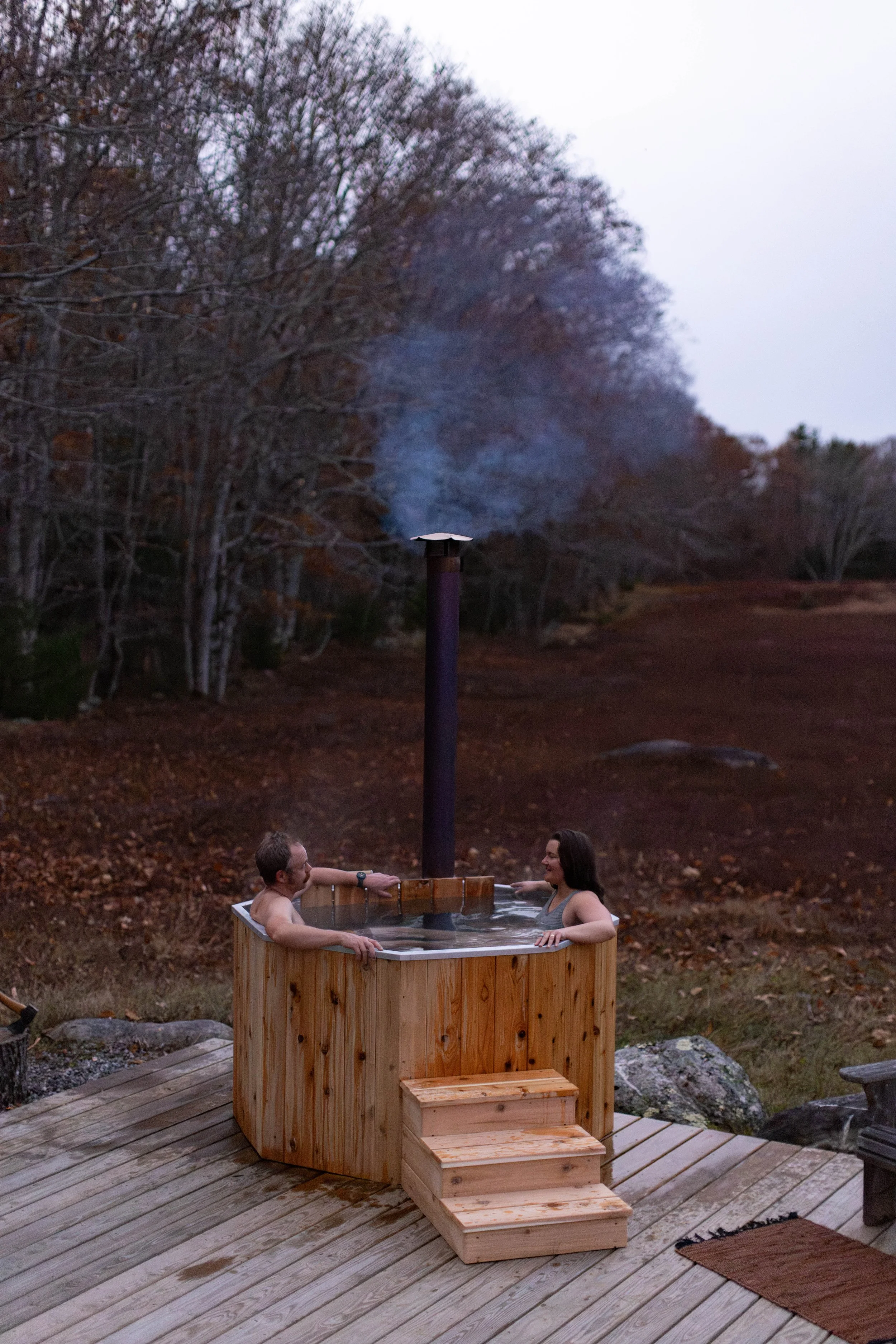 Two people are relaxing in a cedar-clad wood fired hot tub with a chimney emitting steam outdoors during autumn, surrounded by trees with fallen leaves.